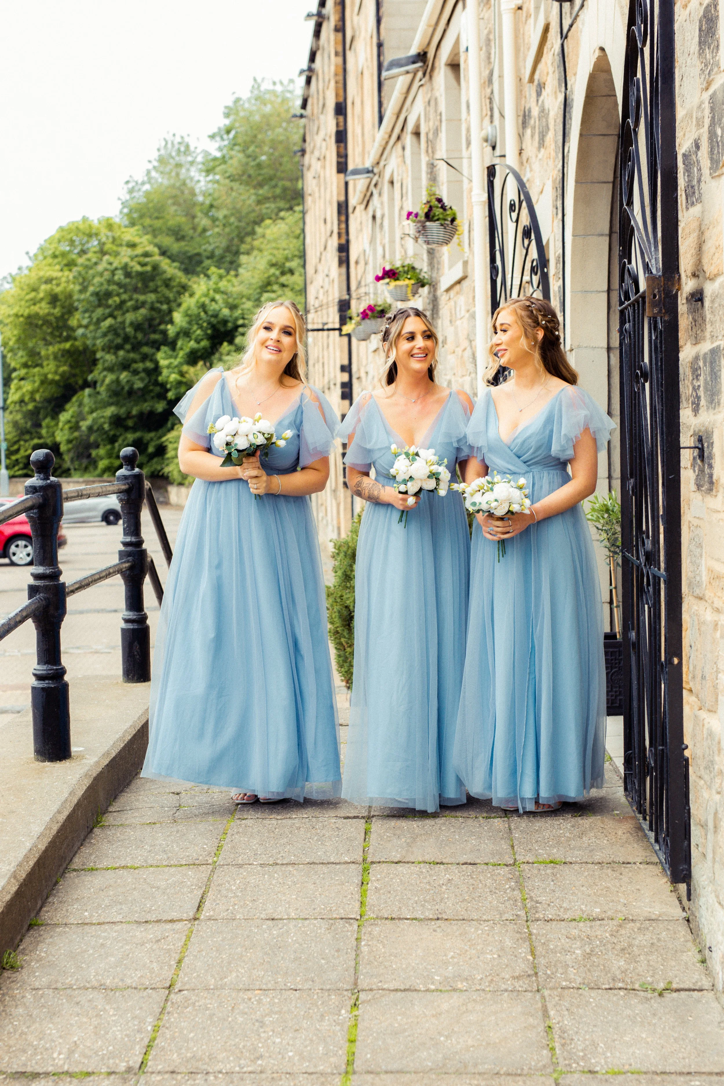 Three bridesmaids in matching light blue dresses holding white bouquets, standing outside on a sidewalk near a stone building with black gates, smiling and interacting with each other.