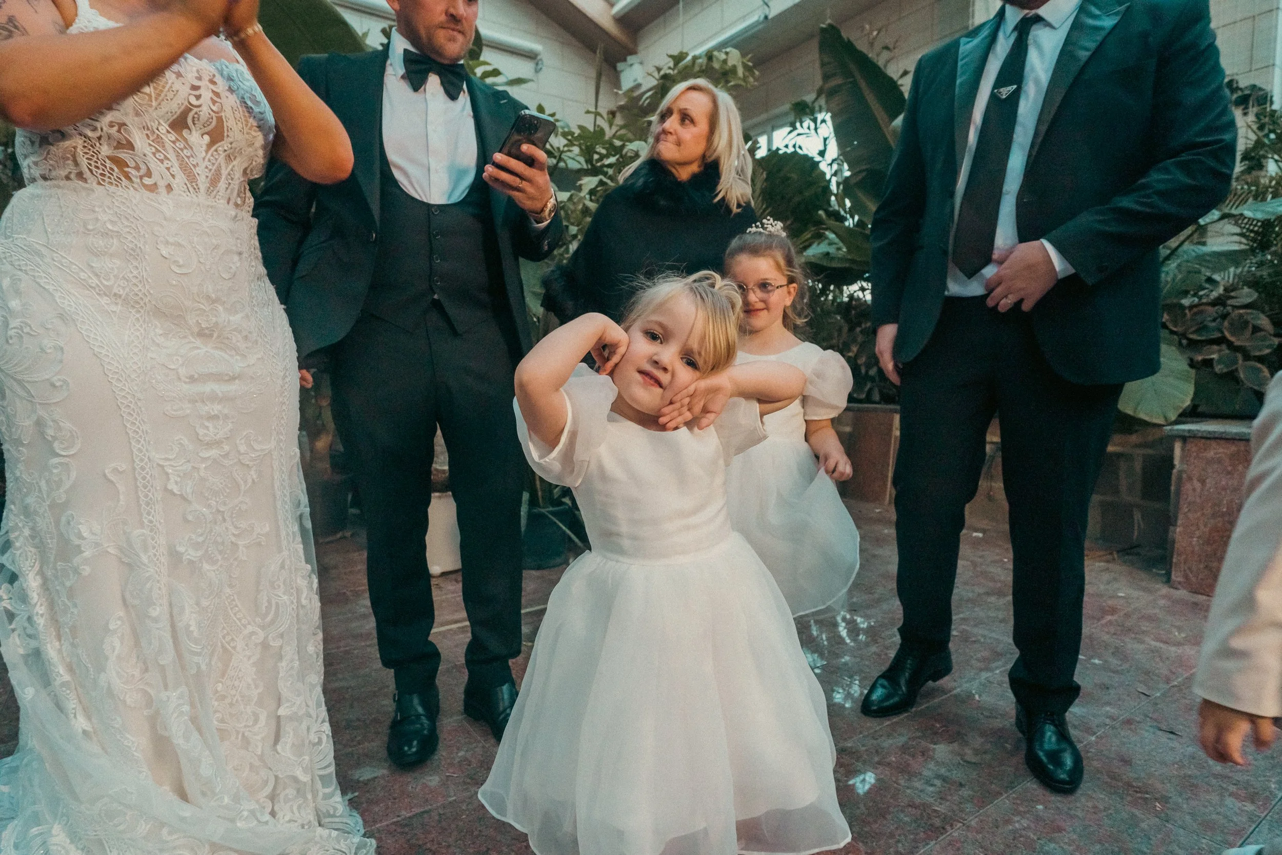Young girl in a white dress making a funny face at a wedding reception surrounded by adults and other children.