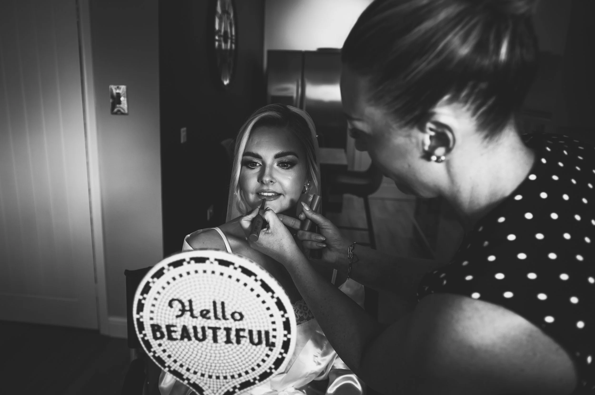 A woman is getting her makeup done by a makeup artist in a room with modern decor. The woman has long blonde hair and is smiling while looking into a mirror. The makeup artist is applying lipstick while holding a lipstick tube, and a sign that reads 