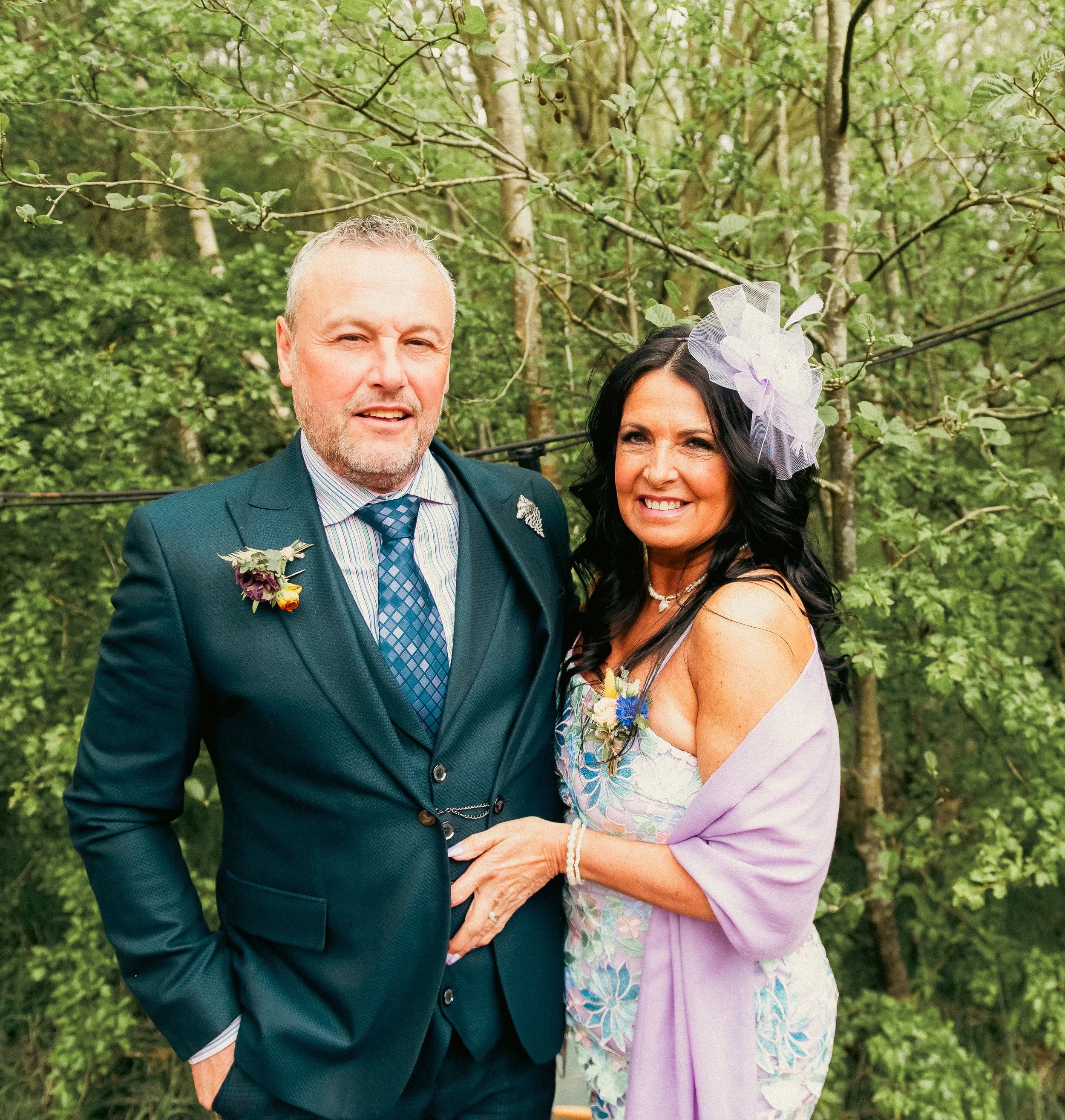 A man and woman dressed in formal attire standing outdoors among green trees, smiling at the camera. The man wears a dark suit with a striped shirt and tie, and the woman wears a colorful dress with a lavender shawl and a large floral hat.