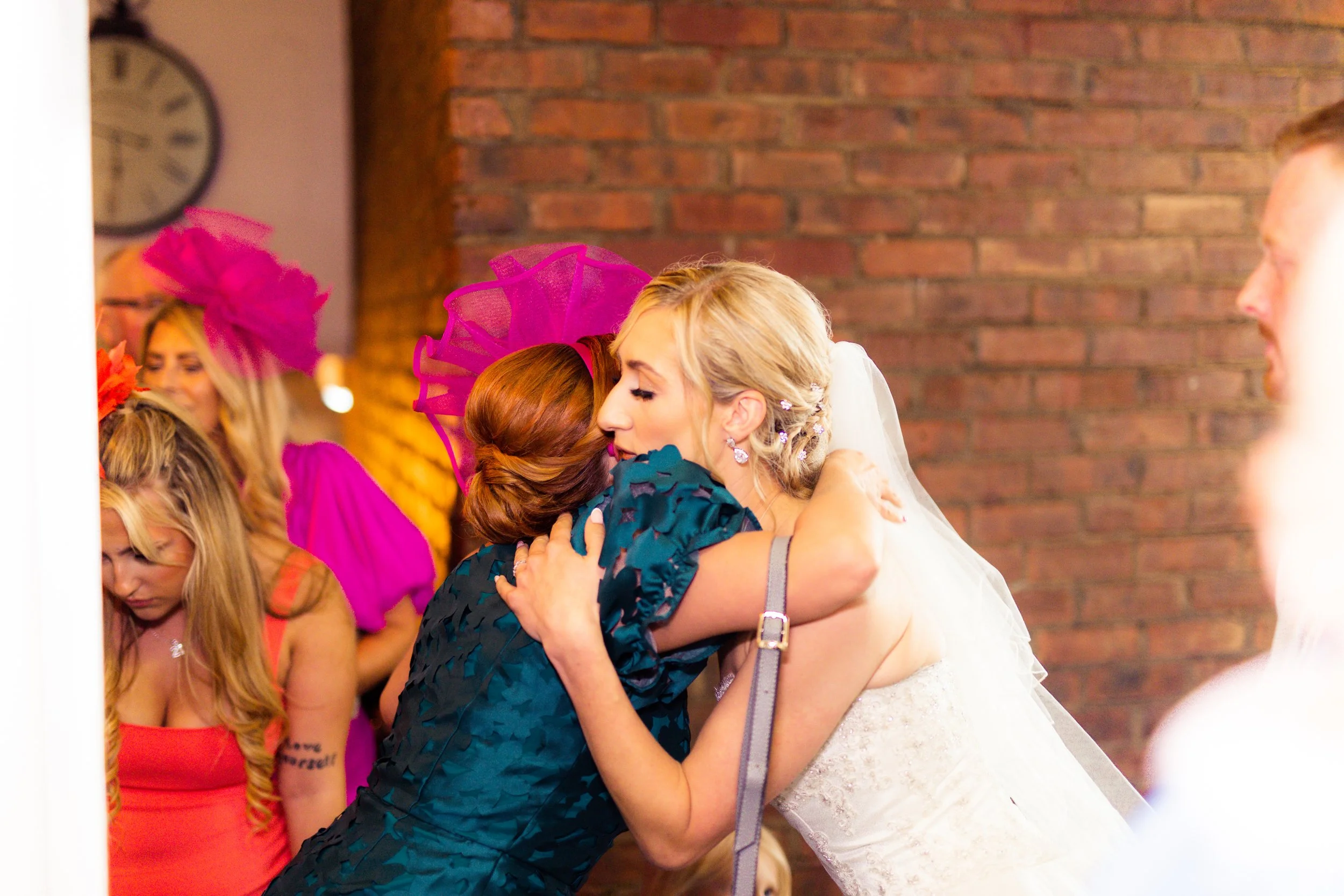 A bride hugging a woman at a wedding reception with other women in colorful hats in the background.