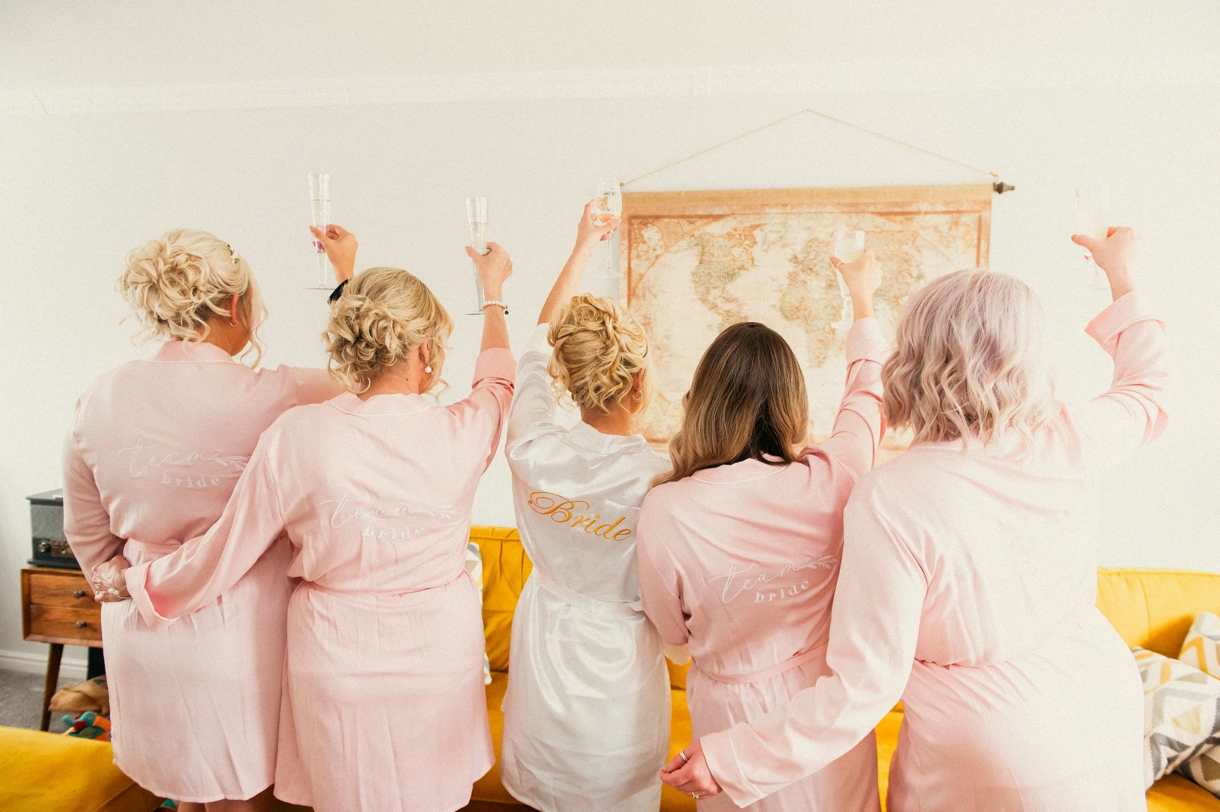 Five women dressed in matching pastel-colored robes, with one robe labeled 'Bride,' celebrate and raise champagne glasses in a living room with a world map on the wall.