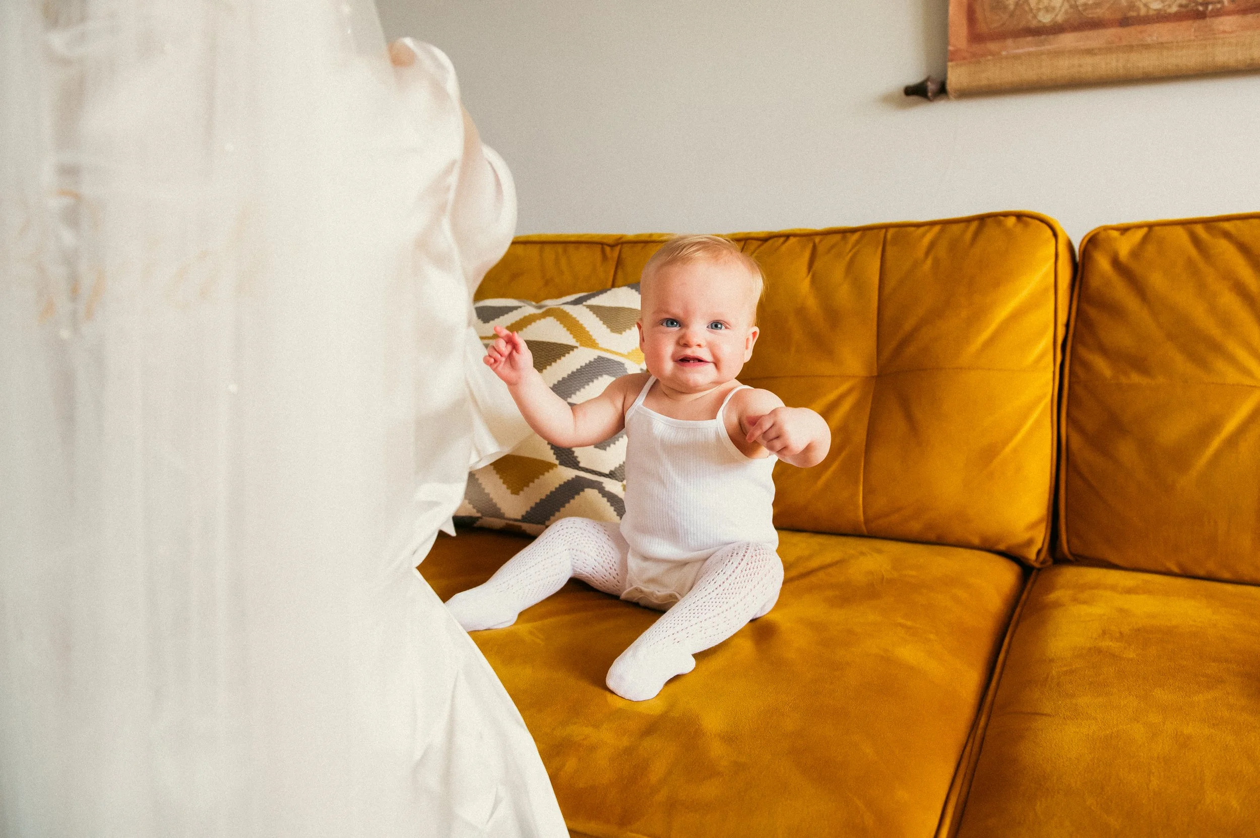 A baby girl with a upset expression, wearing a white tank top and white knit tights, sitting on a mustard yellow couch, reaching towards a person who is partially visible.