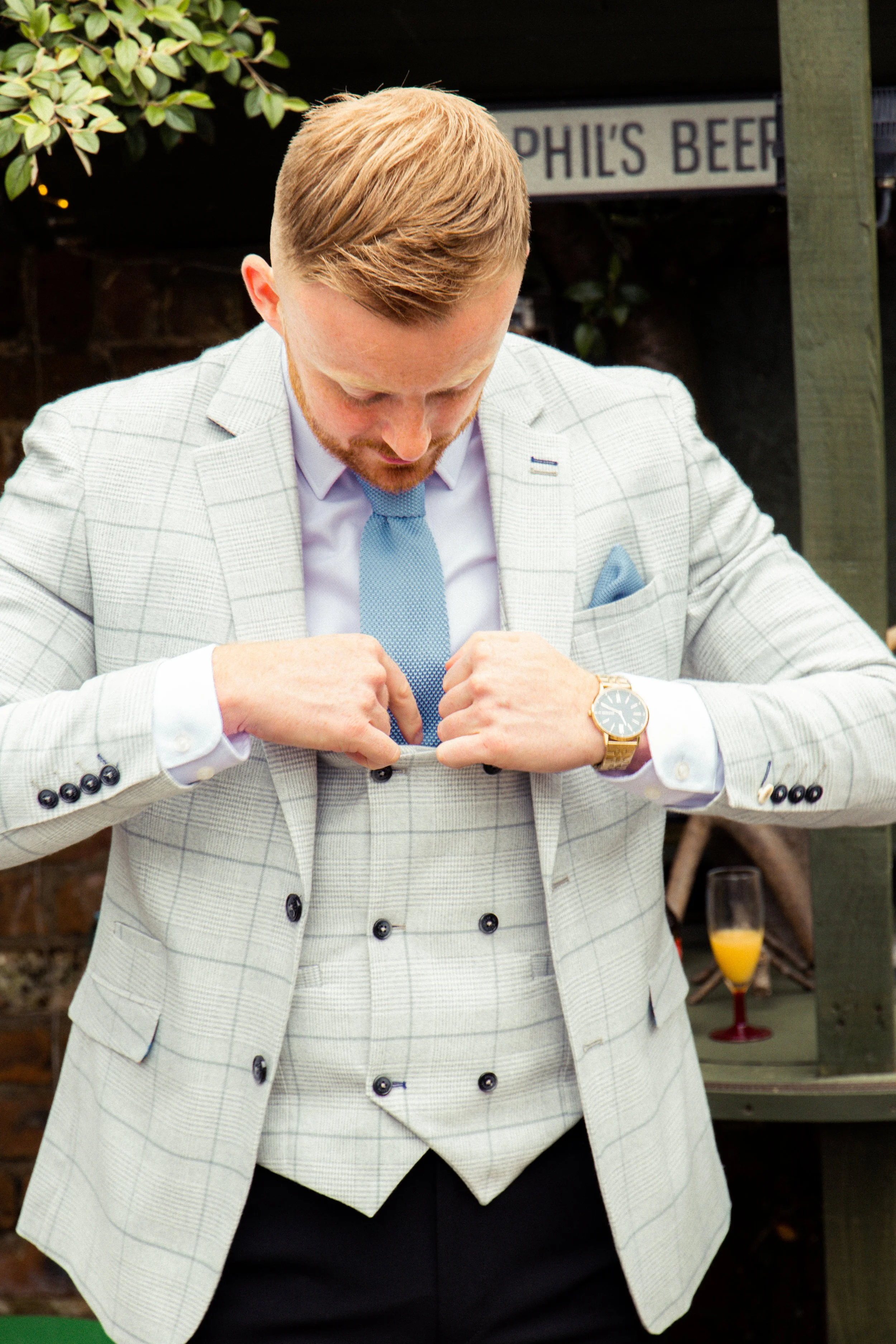 A man in a light gray checkered suit with a blue tie is buttoning his jacket. He is standing outdoors with a sign that reads 'PHIL'S BEER' in the background, alongside some greenery and a glass of orange drink on a table.