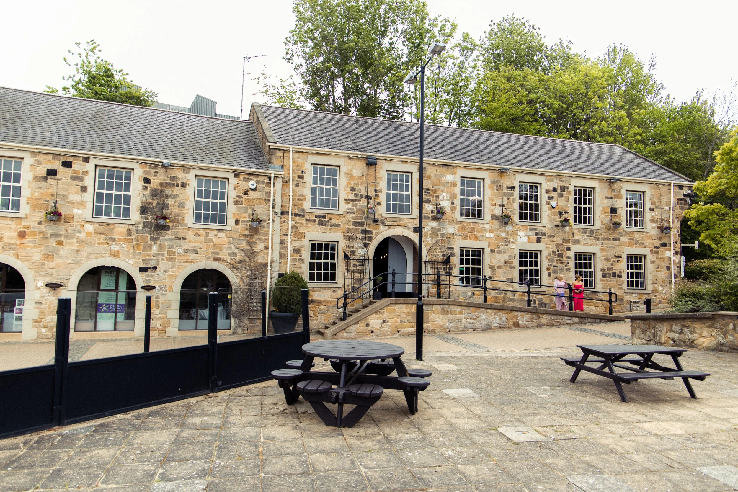 A stone building with multiple windows and flower baskets, featuring a raised entrance with stairs, a black railing, and a small outdoor seating area with picnic tables, two women standing and talking on a paved area.