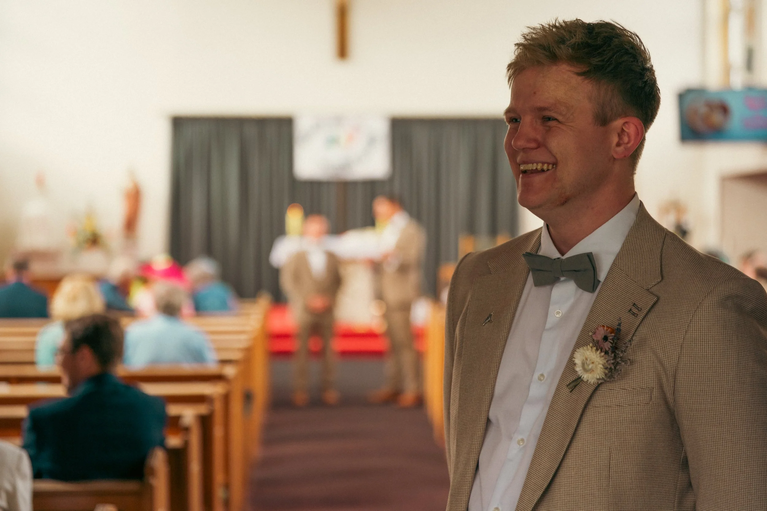 A smiling man in a tan suit with a bow tie, standing at a wedding ceremony, with blurred guests and an altar in the background.
