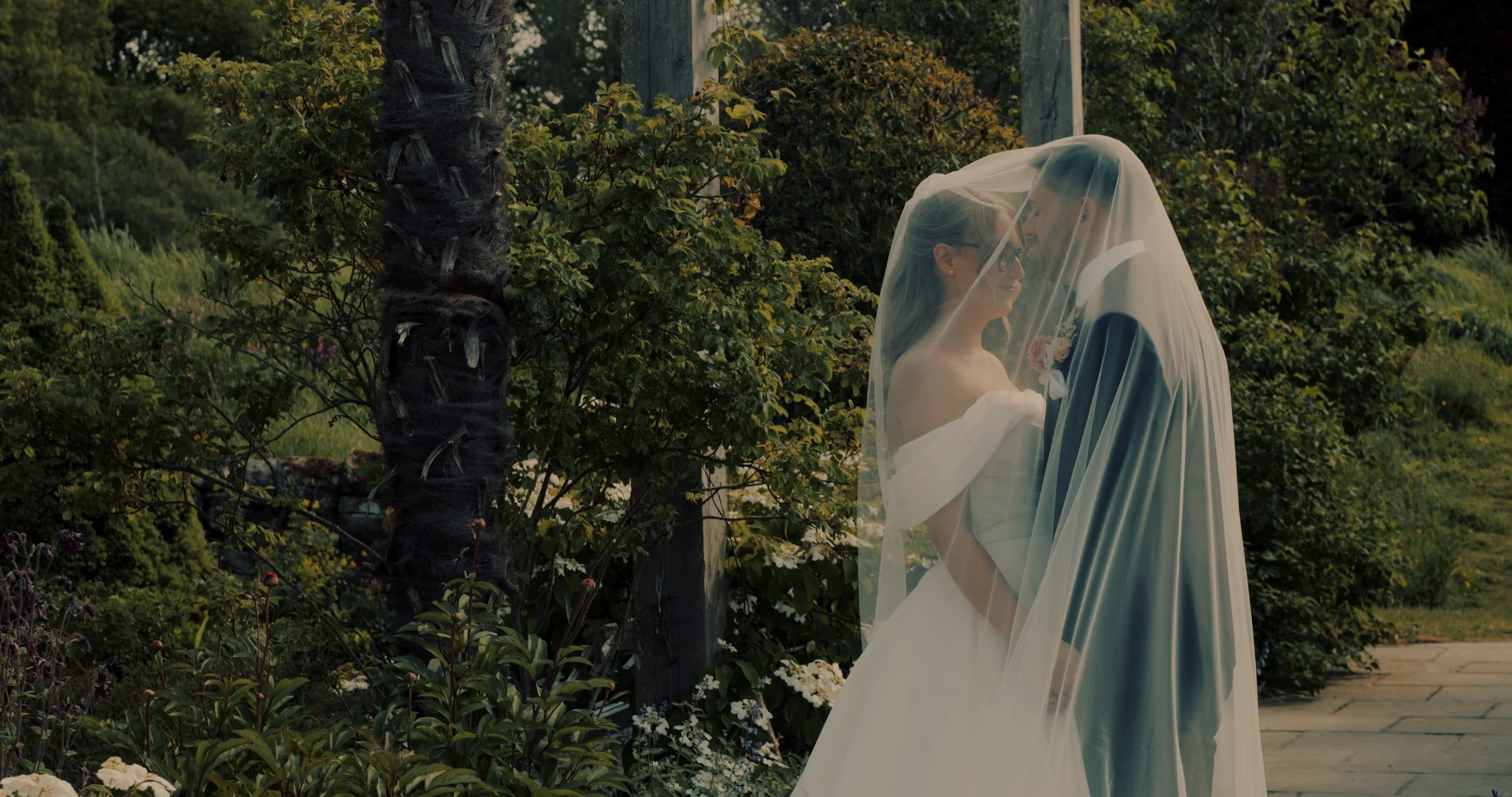 Bride and groom standing close under a veil outdoors, surrounded by greenery and flowers.