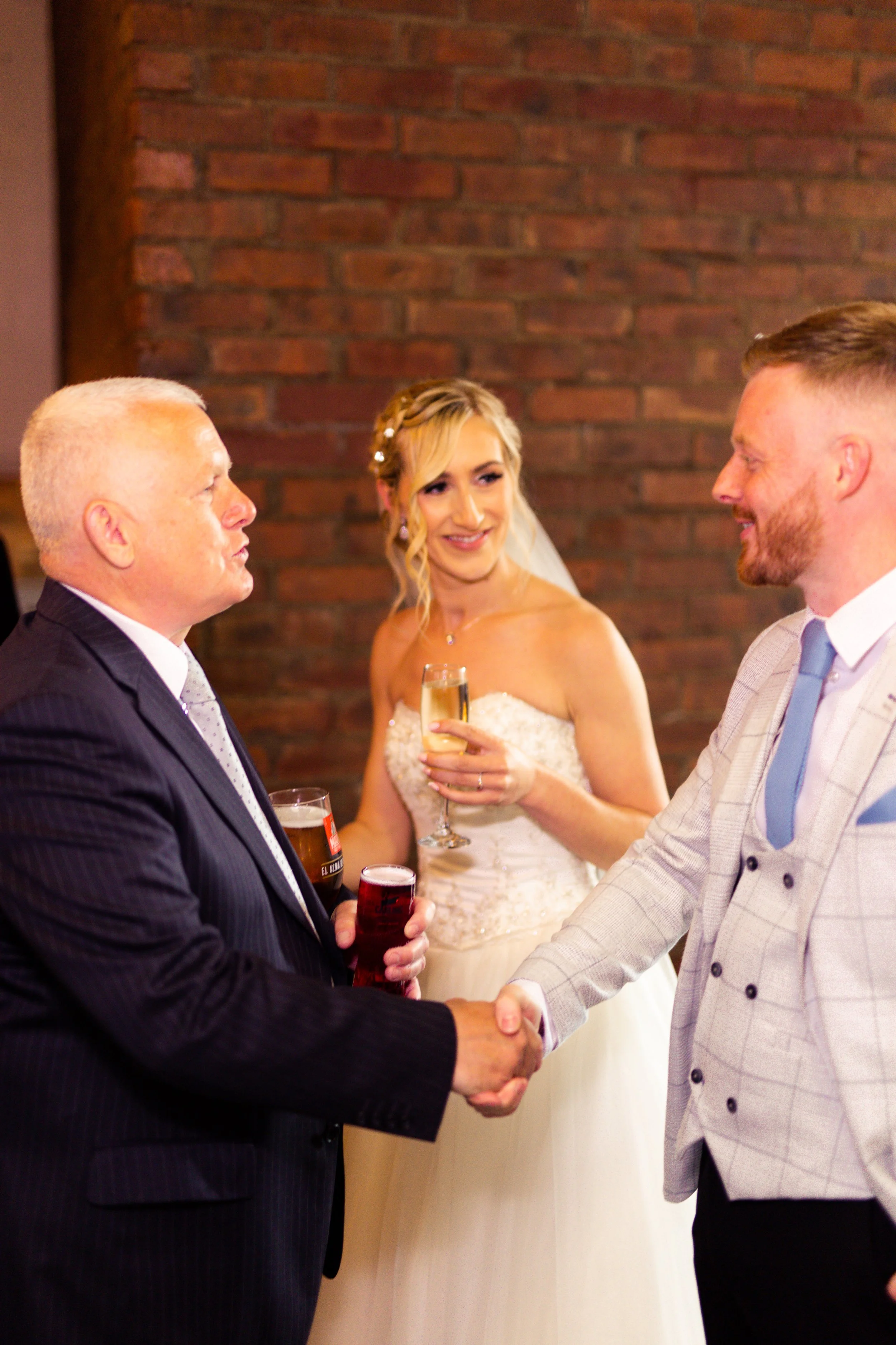 A bride in a wedding dress talking with two men, one in a dark suit and another in a light suit, at a wedding reception, holding glasses of champagne and beer, in front of a brick wall.