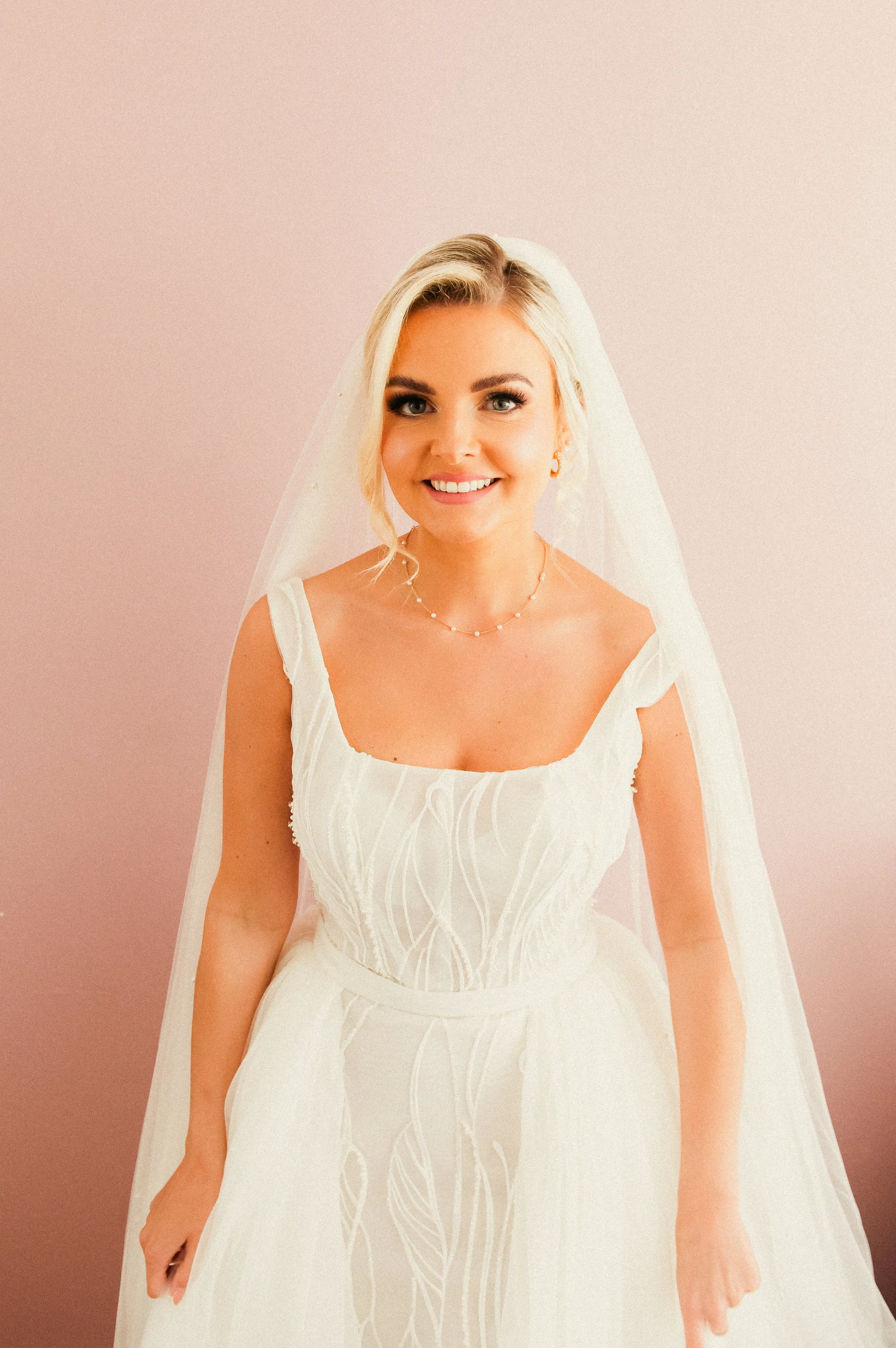 A smiling bride in a white wedding dress with lace details, wearing a pearl necklace and veil, standing against a light pink background.