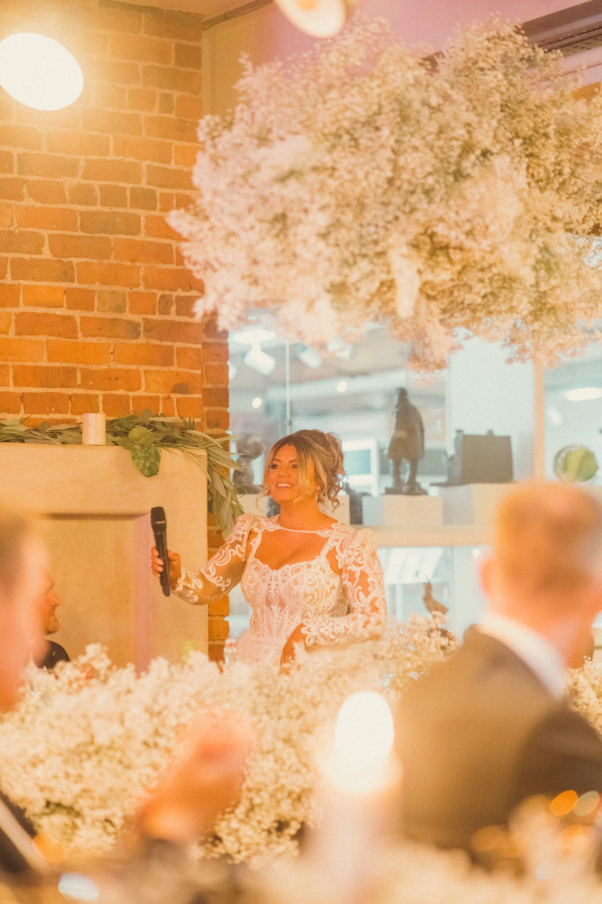 A woman in a white lace dress giving a speech at a wedding reception, with floral decorations and warm lighting.