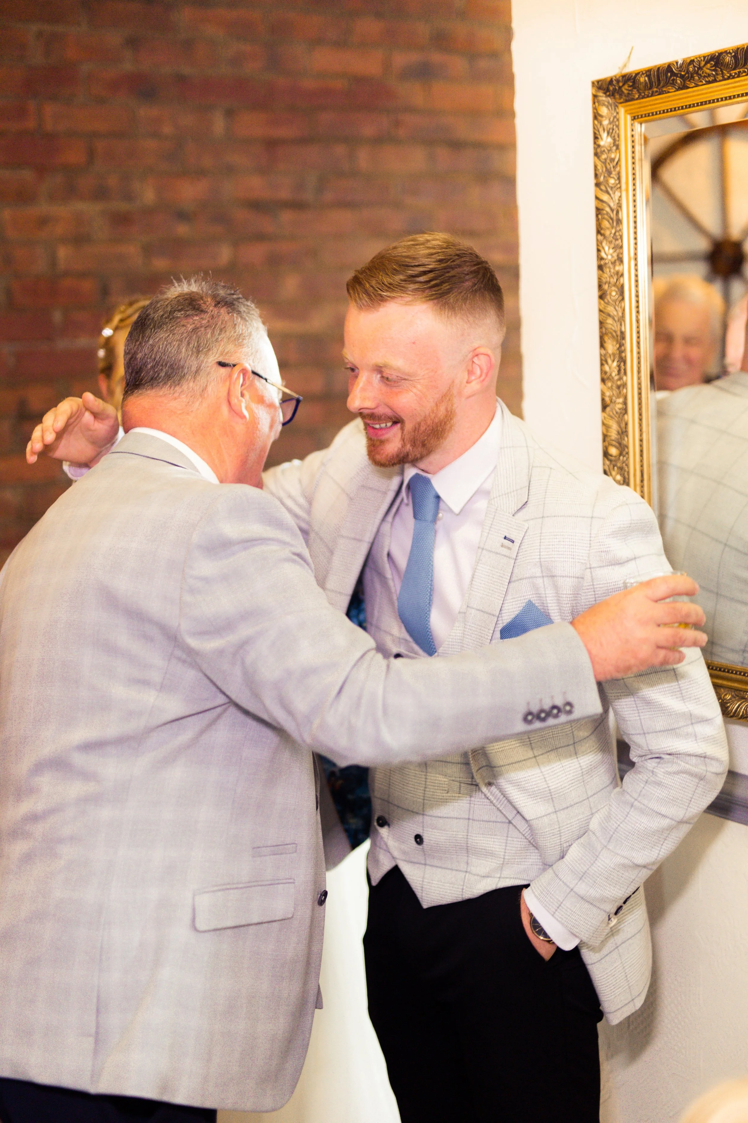 Two men, both wearing light gray plaid suits, are embracing and sharing a joyful moment, with one man smiling warmly at the other. A mirror with an ornate gold frame reflects part of their interaction. The background features a brick wall.