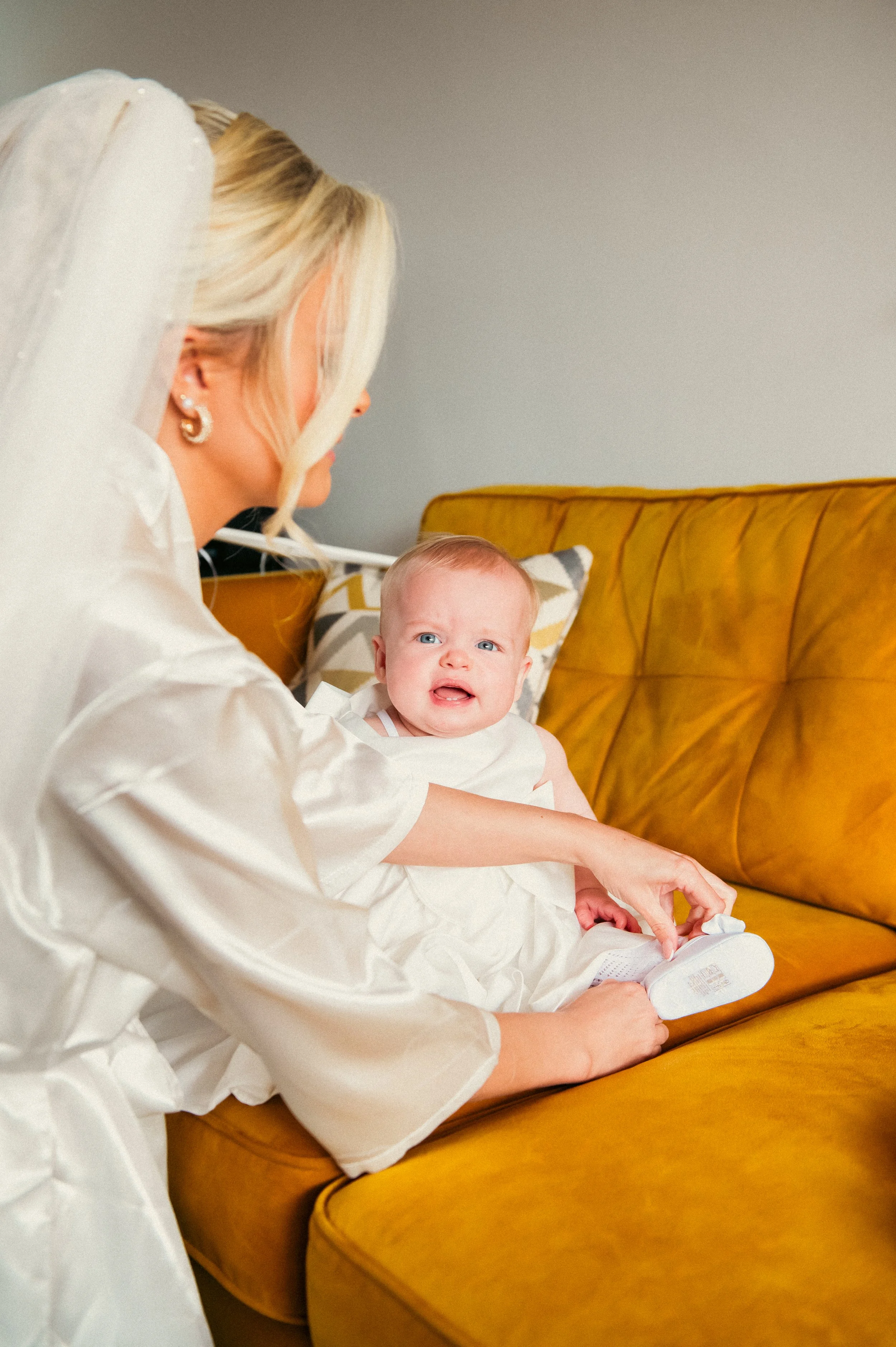 A woman with blonde hair and pearl earrings helping a crying baby with blue eyes put on a white shoe on a yellow couch.