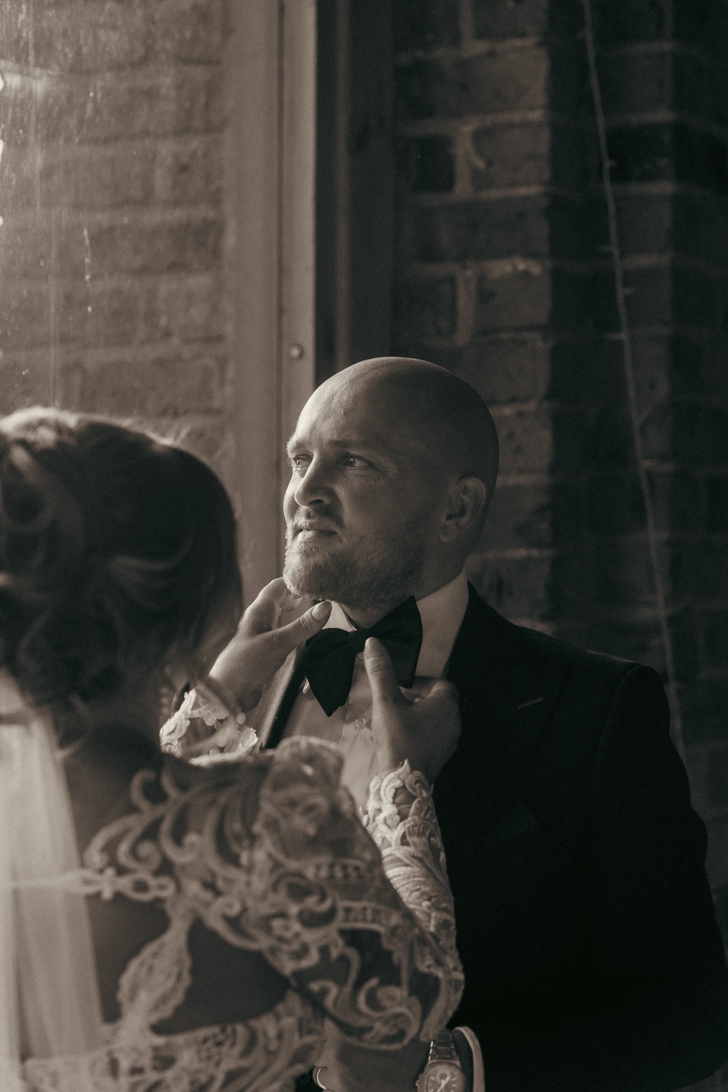 A woman helping a man adjust his tuxedo bow tie, with a brick wall background.