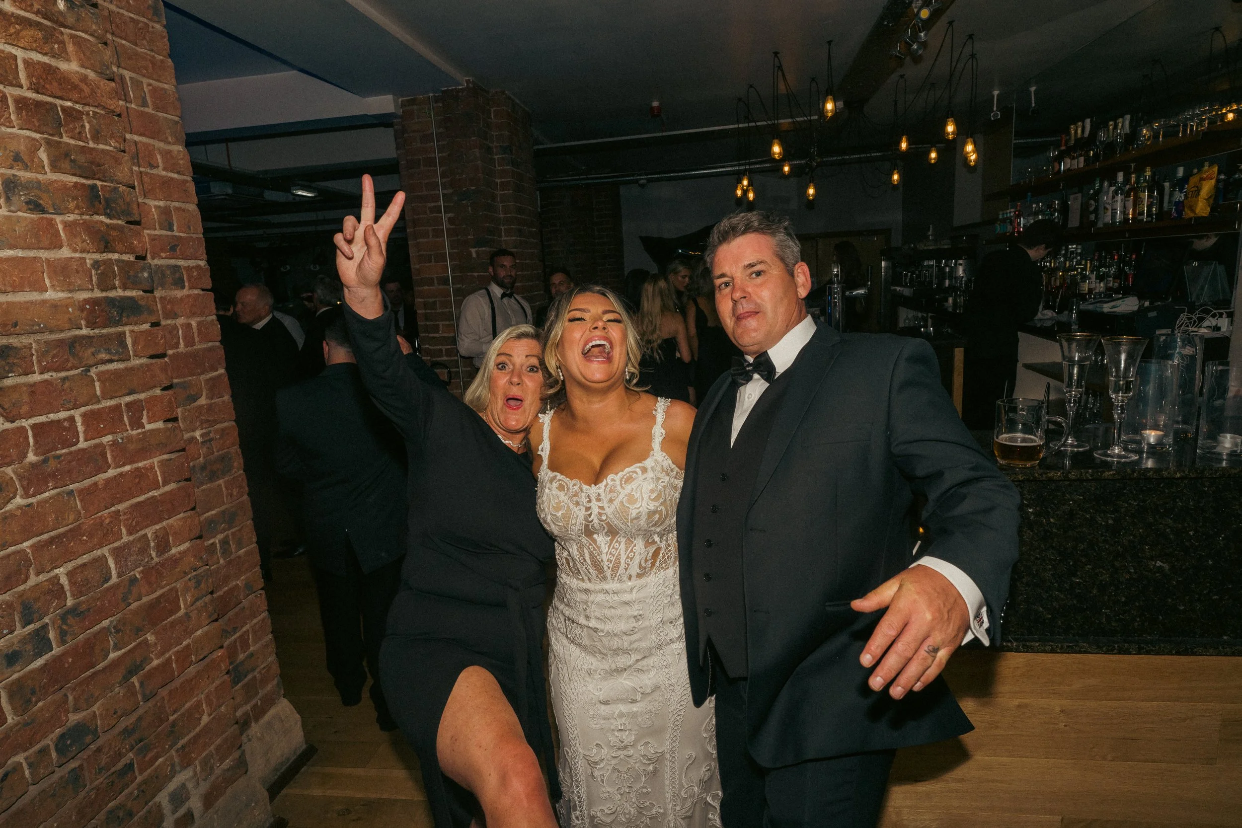 Three people at a wedding reception, a woman in a white wedding dress, a woman in a black dress with a high slit, and a man in a tuxedo, posing playfully for the camera.