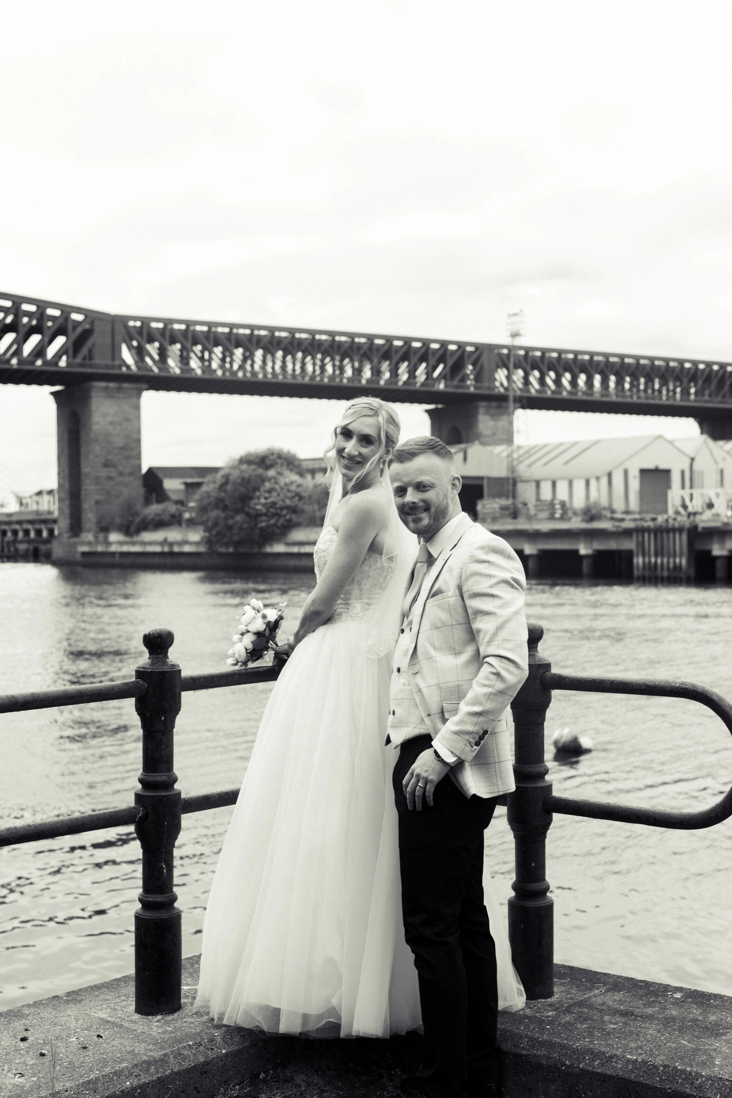 A black-and-white photo of a bride and groom smiling by a waterfront with a bridge in the background. The bride wears a wedding dress and holds a bouquet, while the groom wears a light-colored suit.