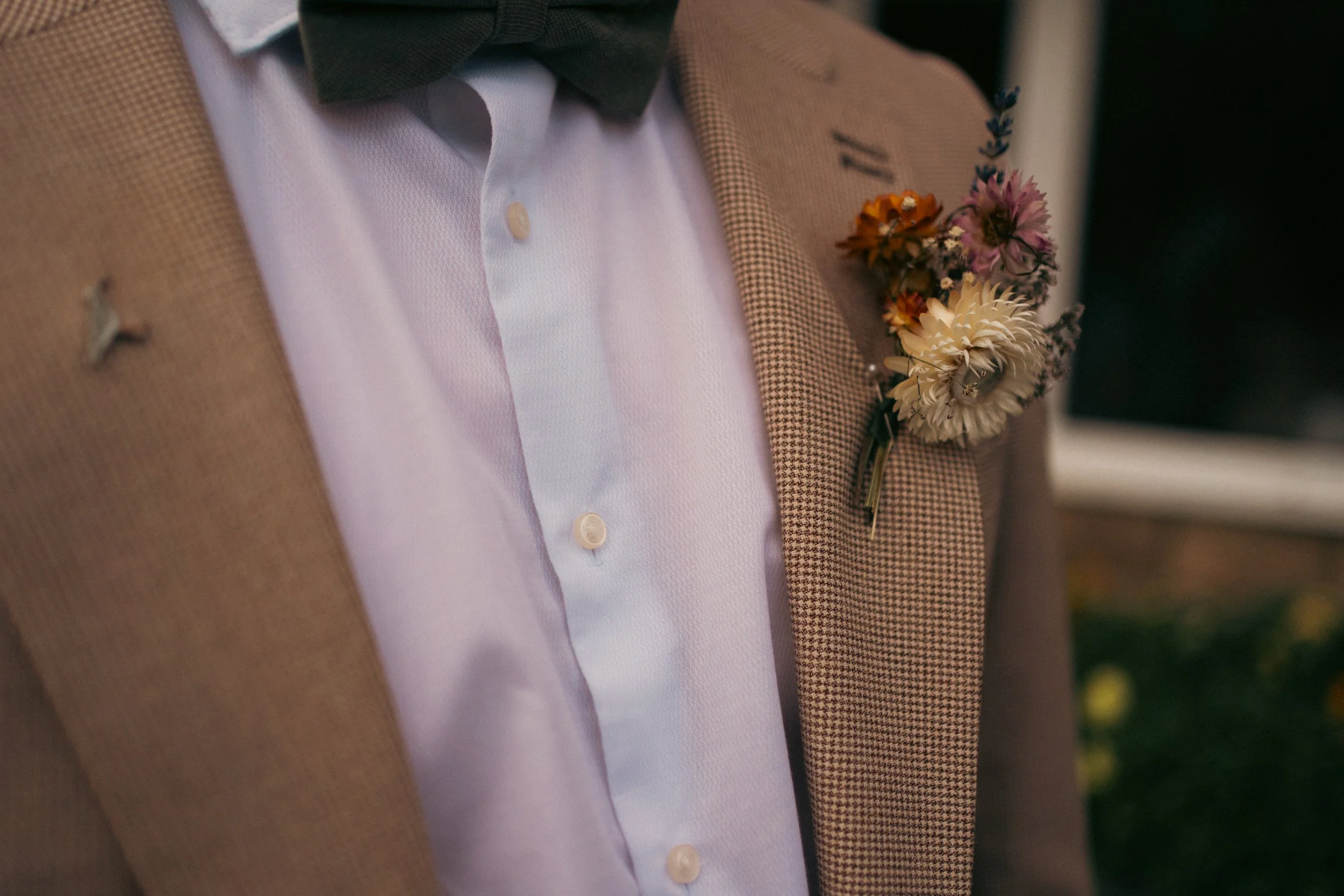 Close-up of a man wearing a light brown checked blazer, white shirt, dark green bow tie, and a flower boutonniere on the blazer's lapel.