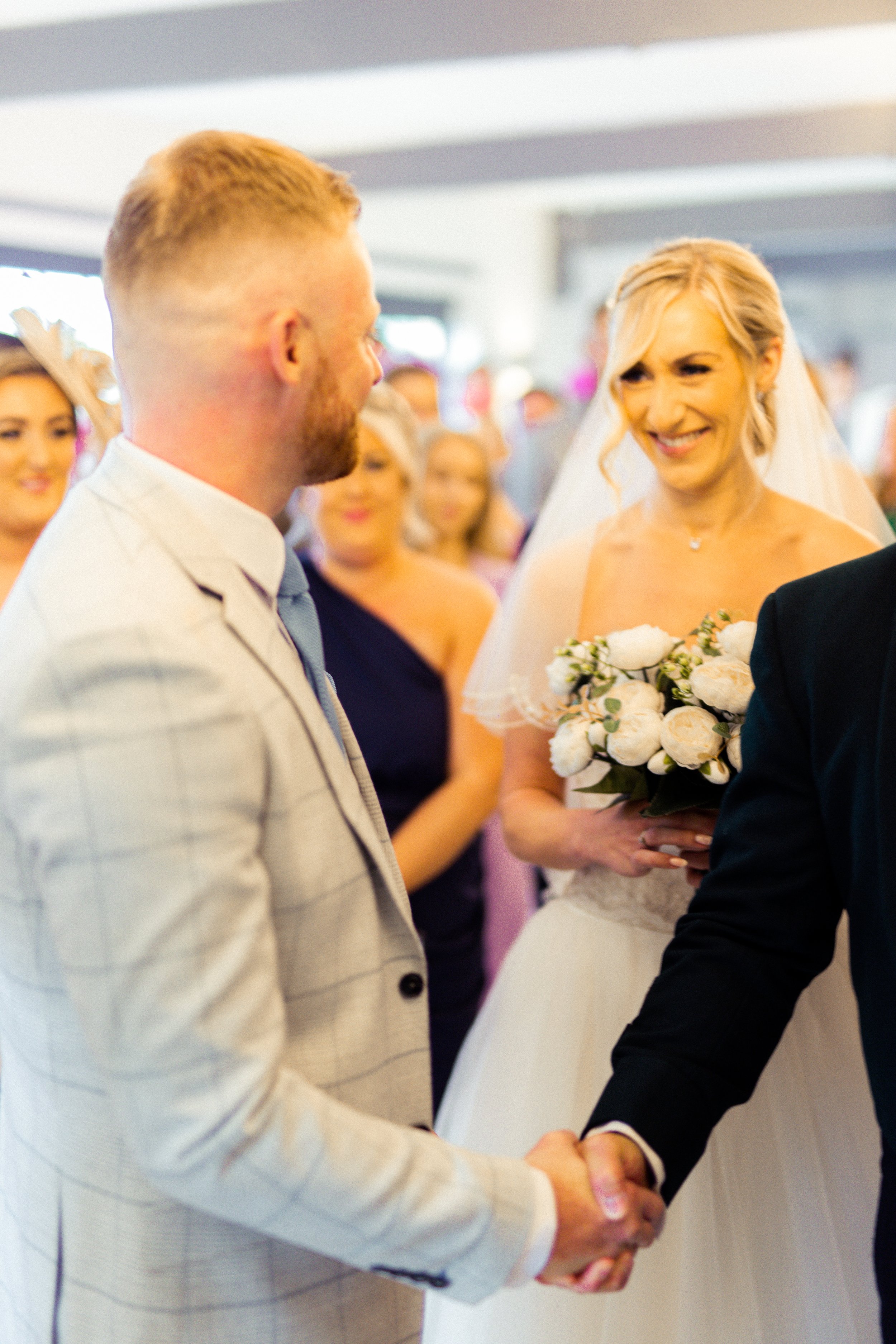A bride and groom shake hands during their wedding ceremony, while a woman in a dark dress looks on, surrounded by guests.