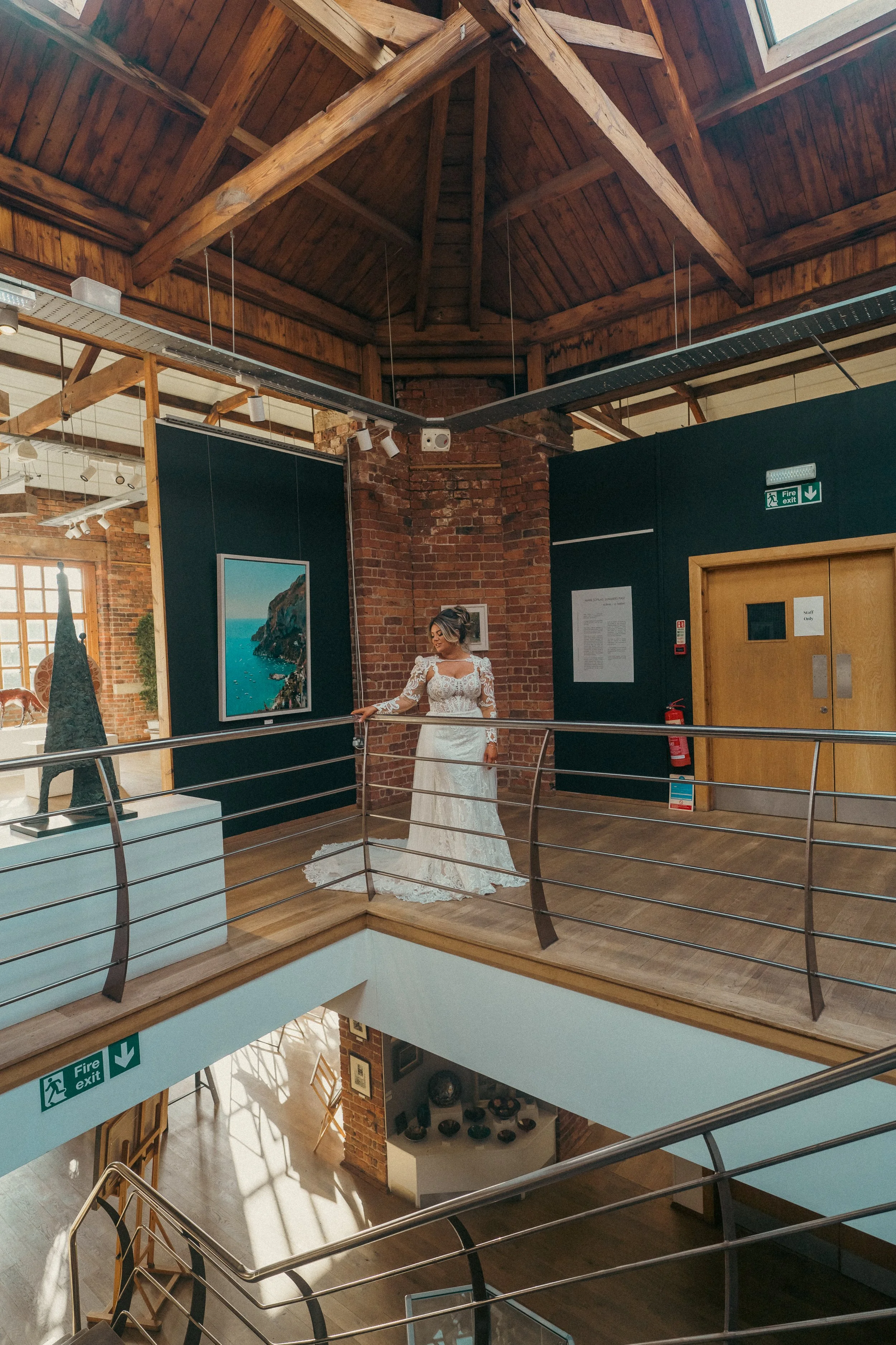 A woman in a white wedding dress standing on a wooden gallery inside a building with brick walls and wooden ceiling beams, looking at artwork on display.