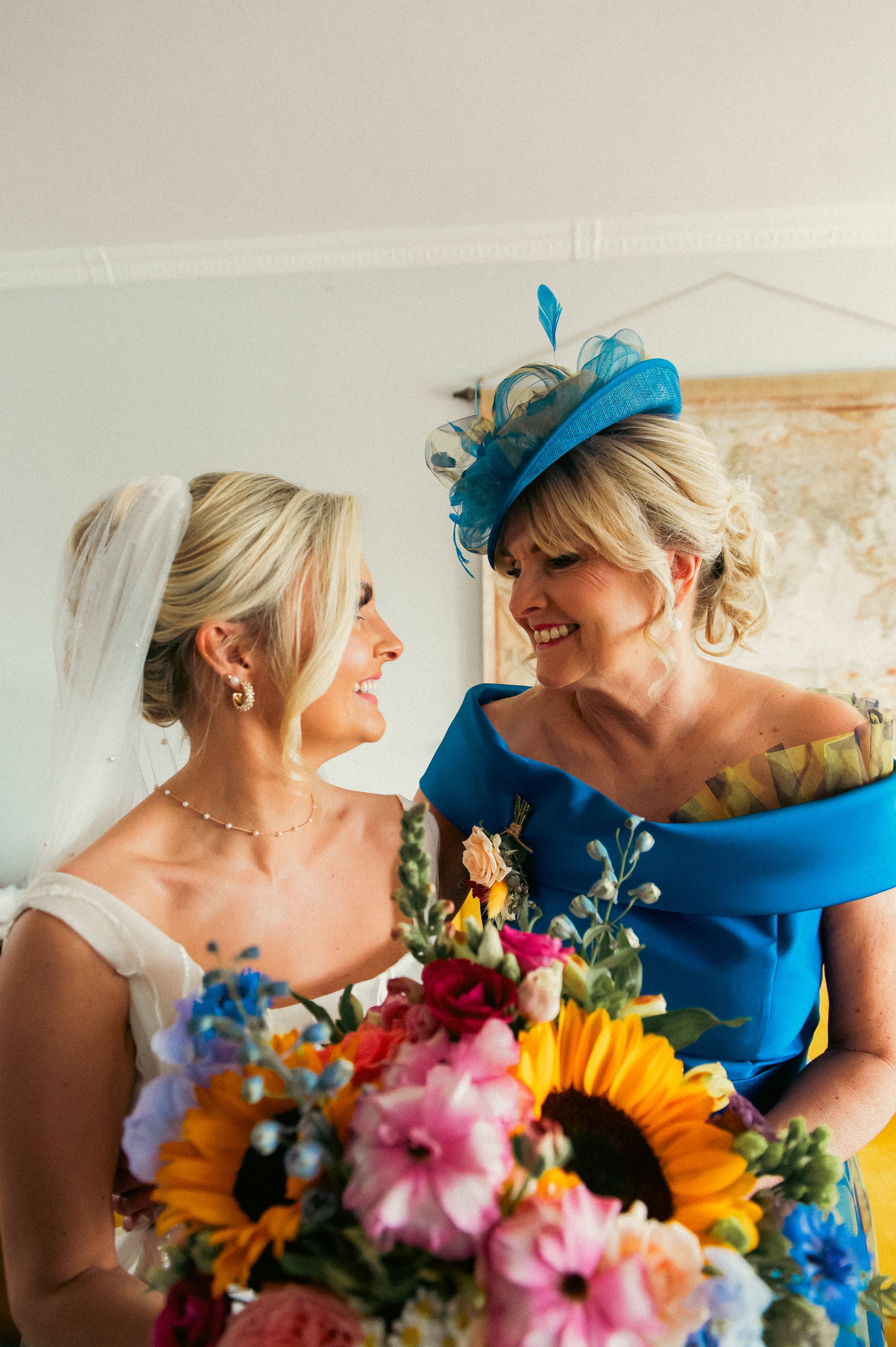 A bride and an older woman smiling at each other, holding a bouquet of colorful flowers. The bride wears a white dress and veil, and the older woman wears a bright blue dress with a matching hat.