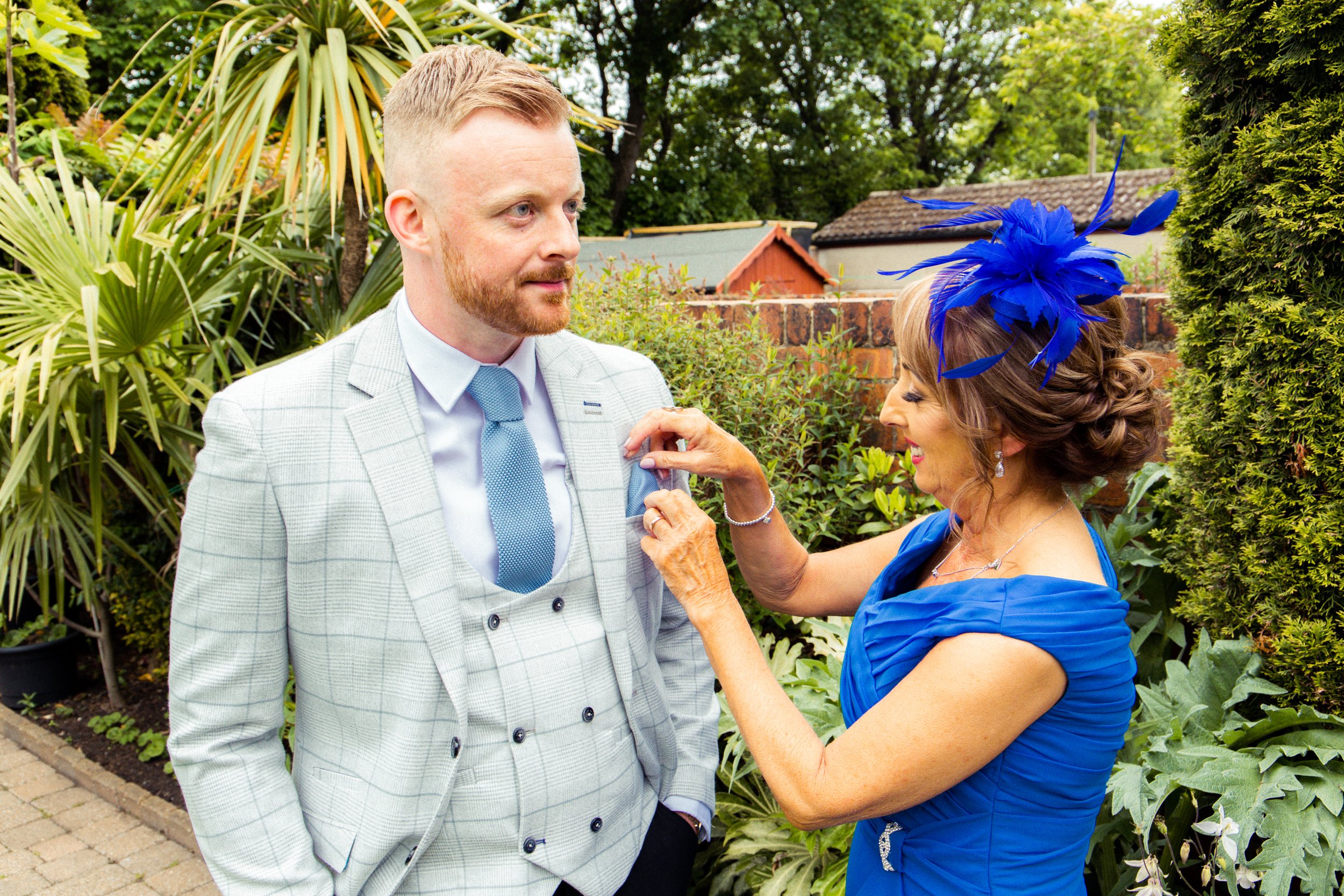 A woman in a blue dress and feathered headpiece pins something to a man's suit jacket outside in a garden.