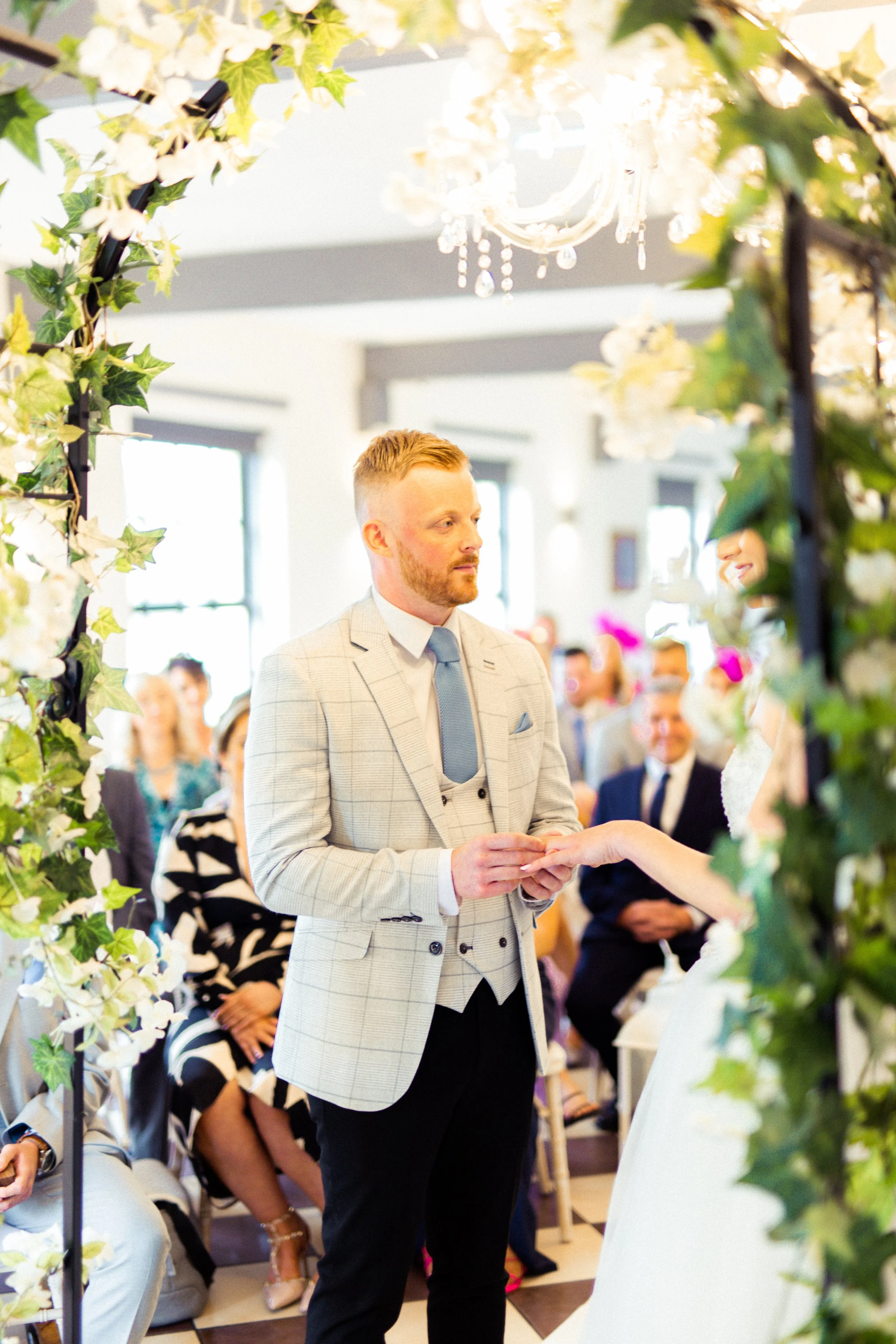 A groom is reciting vows at his wedding ceremony, standing under a floral arch, with seated guests and a chandelier visible in the background.