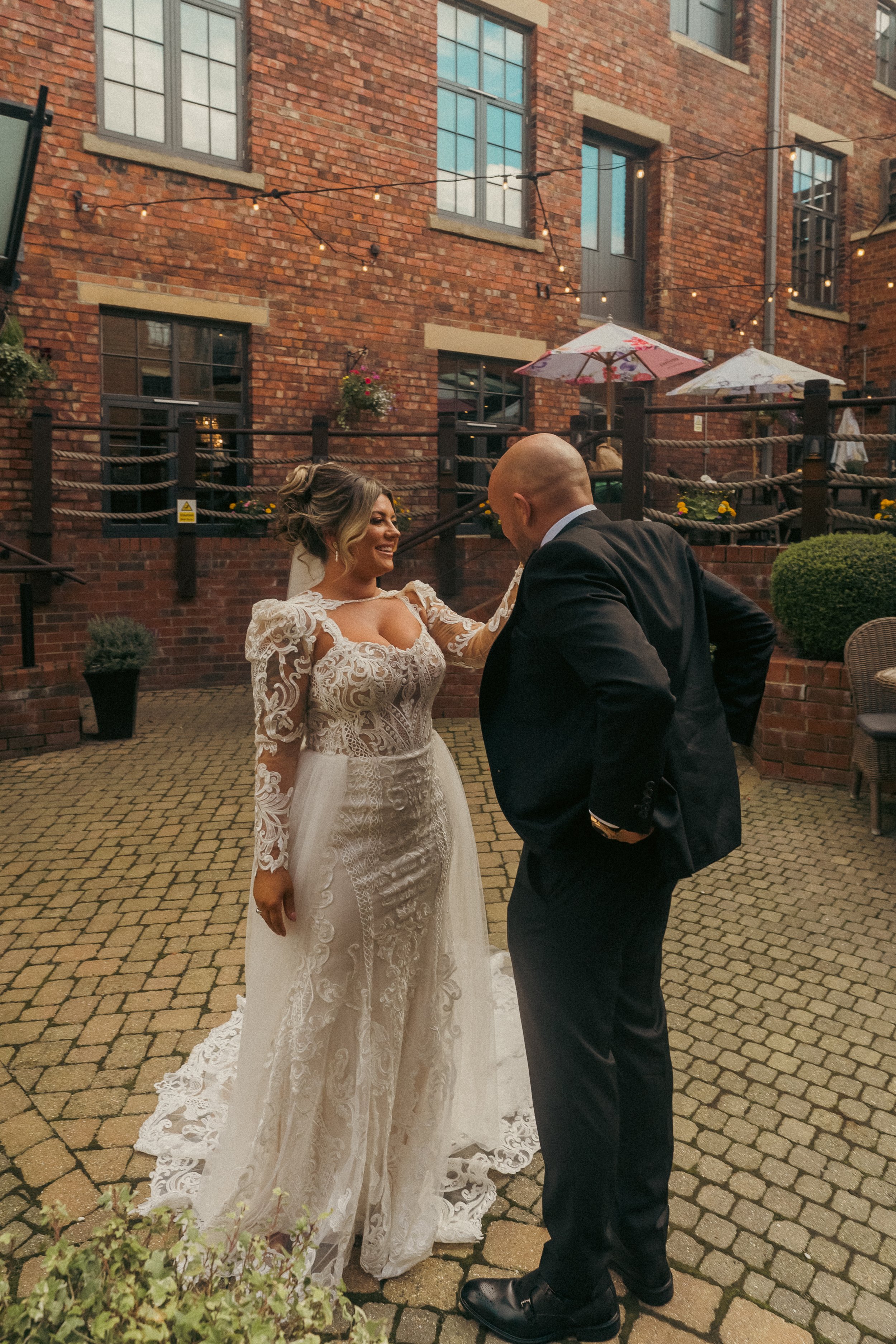 A bride in a lace wedding dress and a groom in a black suit share a joyful moment outdoors on a brick patio, with string lights, umbrellas, and brick buildings in the background.