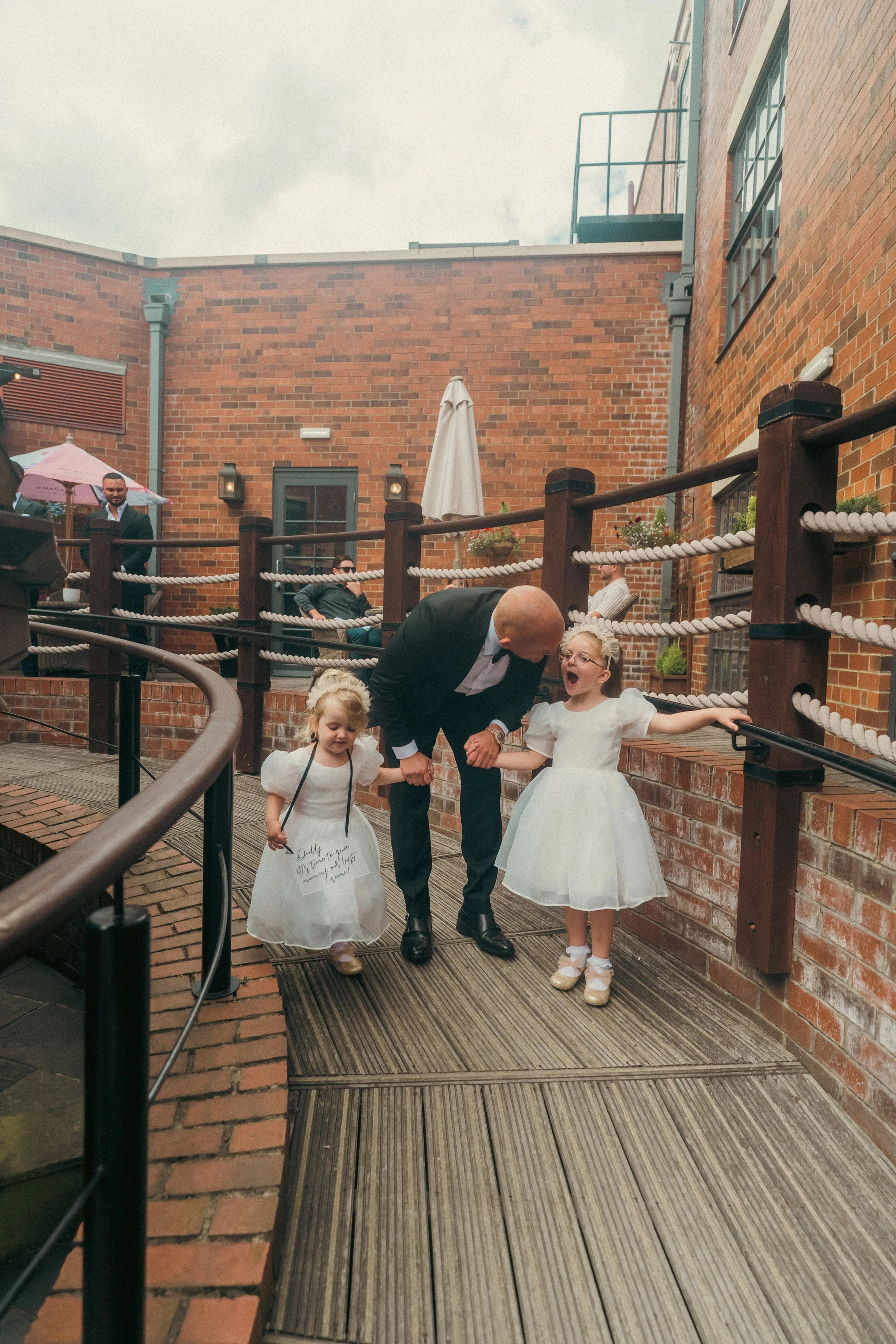 A man in a suit holding hands with two young girls in white dresses, walking down an outdoor brick pathway with a brick wall background, during a celebration on a rainy day.