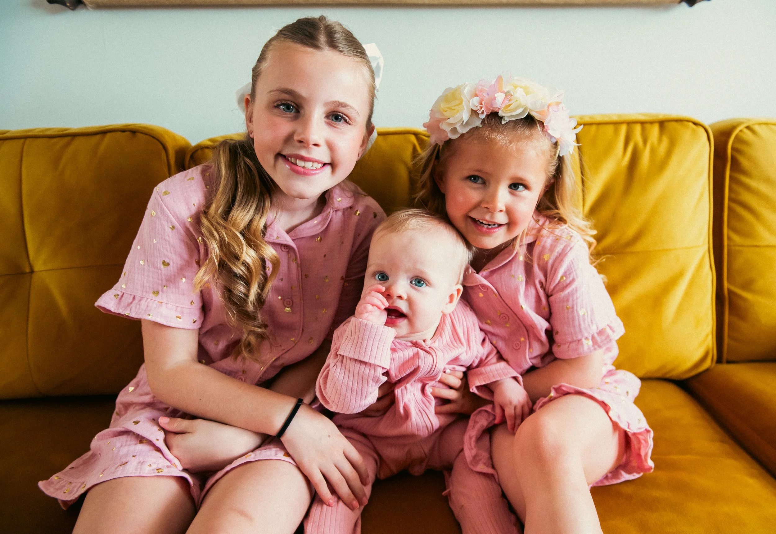 Three young girls sitting on a yellow sofa, smiling at the camera. Two are older with long hair, wearing pink matching outfits with small gold details. The youngest girl, in the middle, has blond hair, blue eyes, and is dressed in a light pink outfit