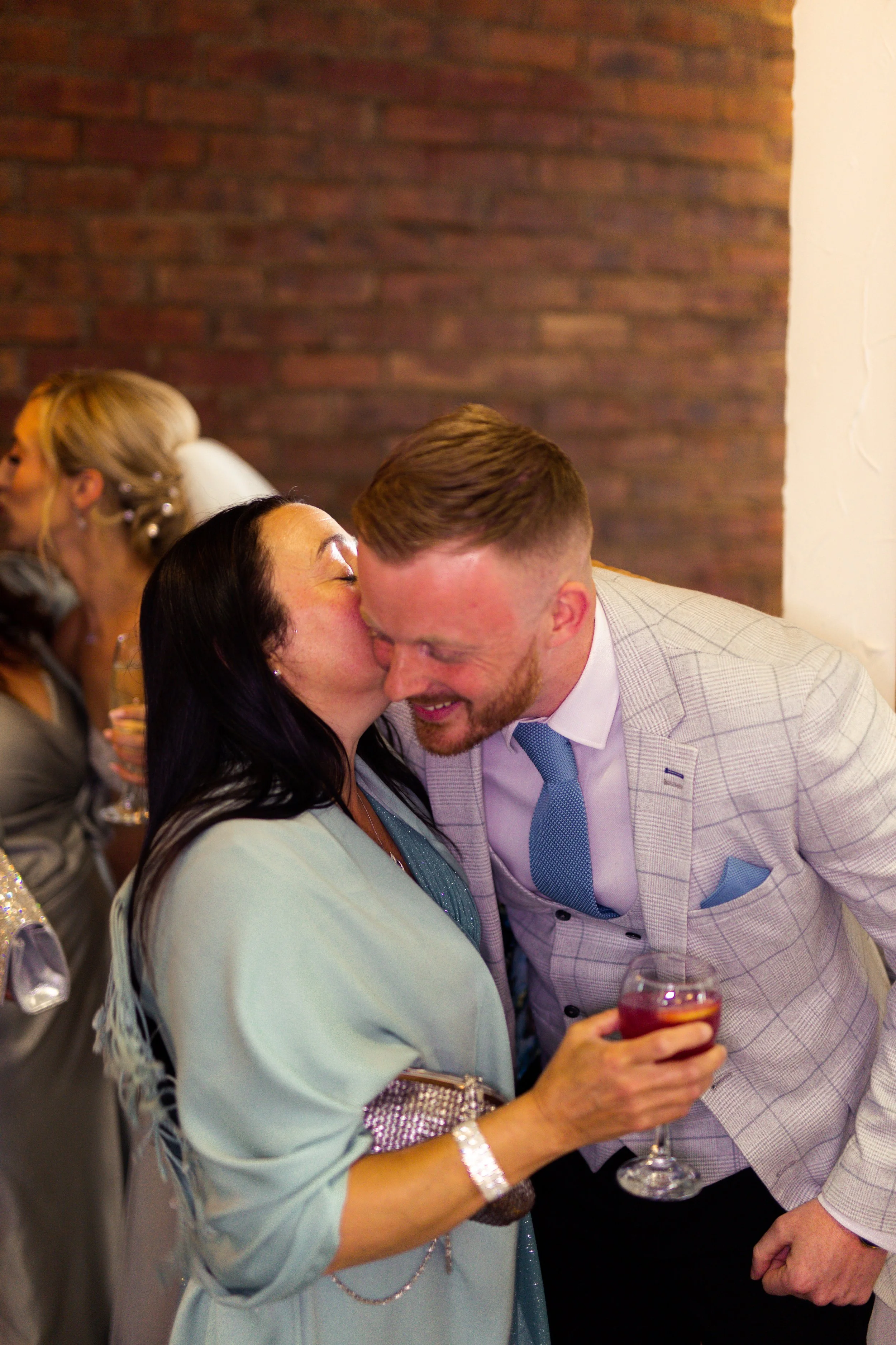 A woman in light blue clothing kissing a man in a light-colored suit on the cheek at a celebratory event, with others in the background celebrating and holding drinks.