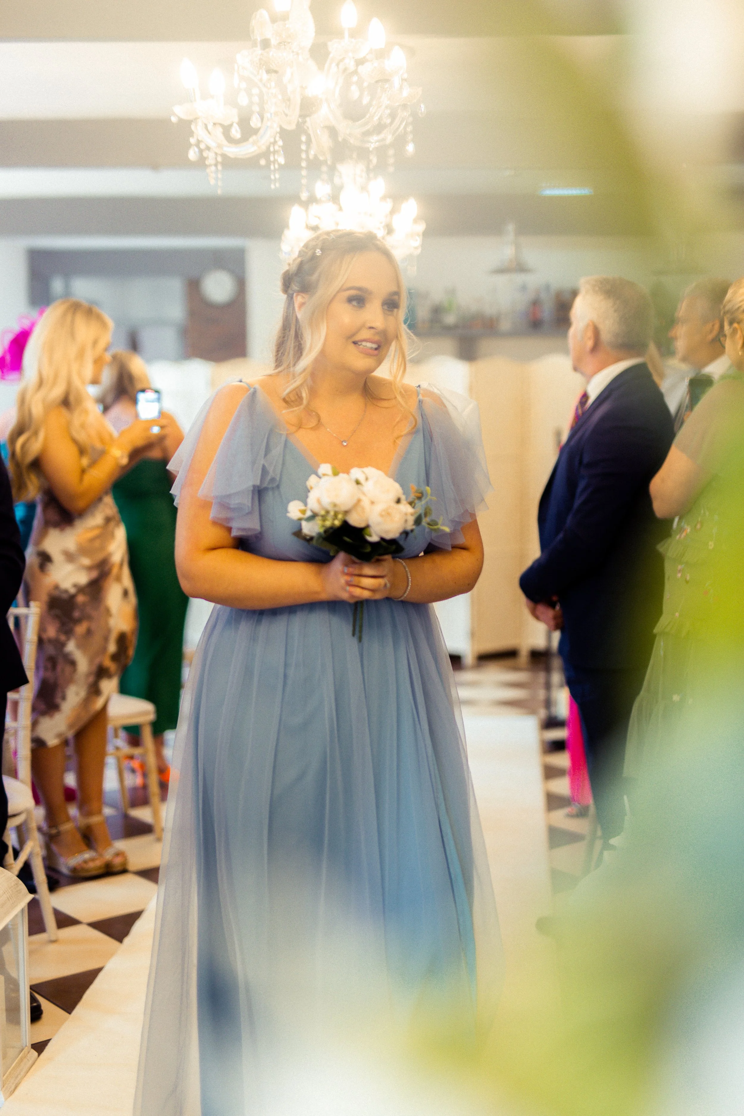 A woman in a light blue dress holding a bouquet of white and pink roses, standing in a decorated room during a wedding or formal event with chandeliers and guests in the background.
