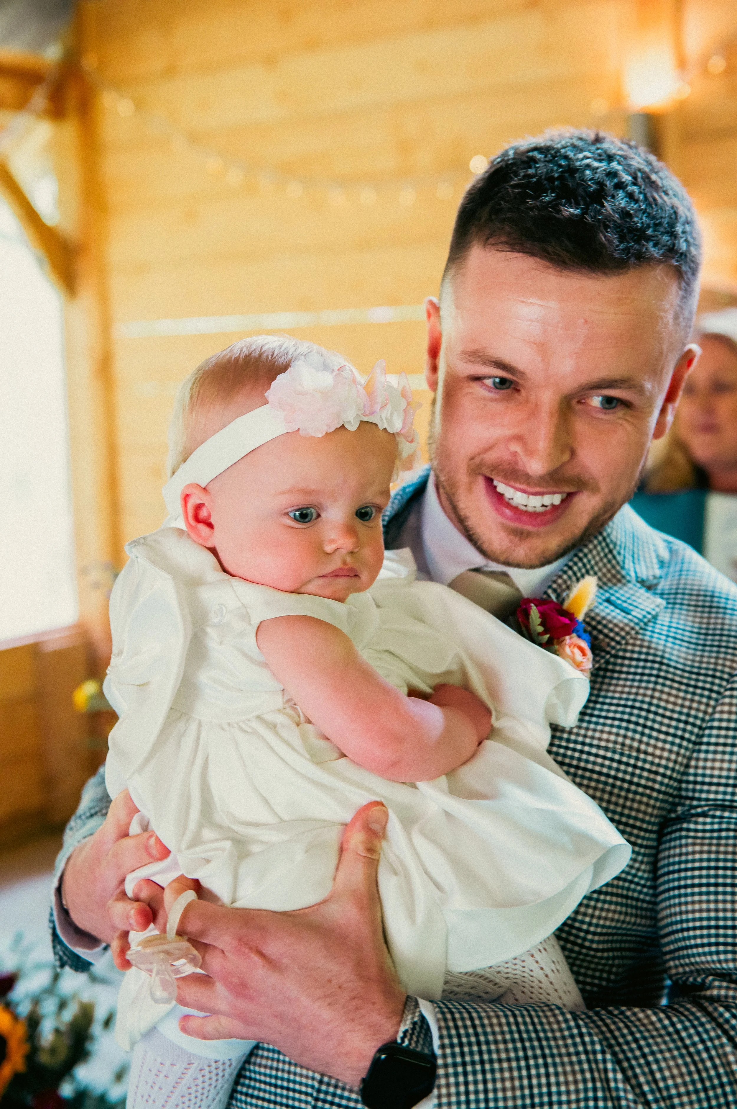 A man with a short beard, dressed in a checkered jacket and bow tie, holding a young girl in a white dress with a floral headband, inside a wooden-paneled room.