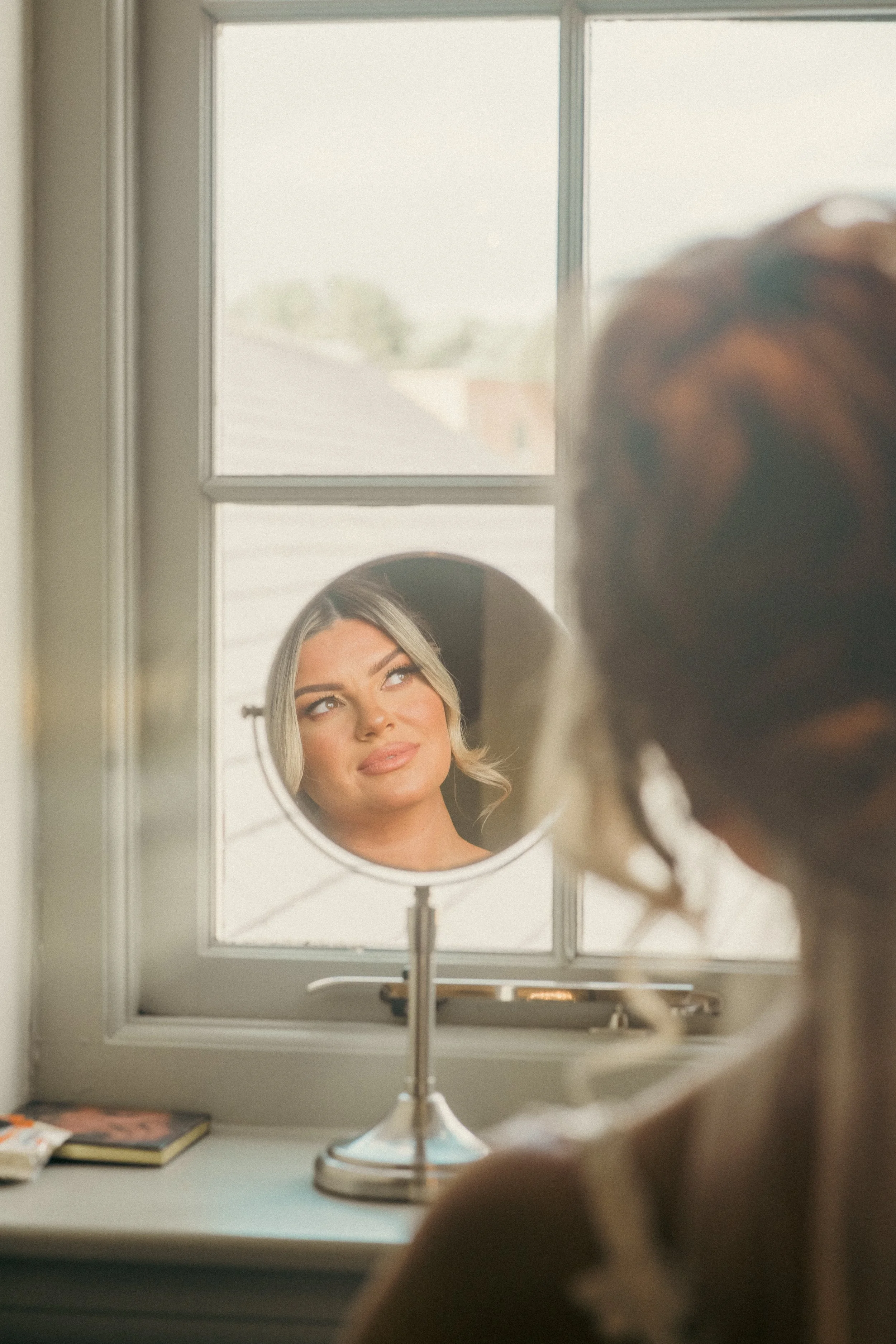 A woman looking into a mirror on a window ledge, with her reflection showing her face and makeup, while the back of her head is blurred in the foreground.