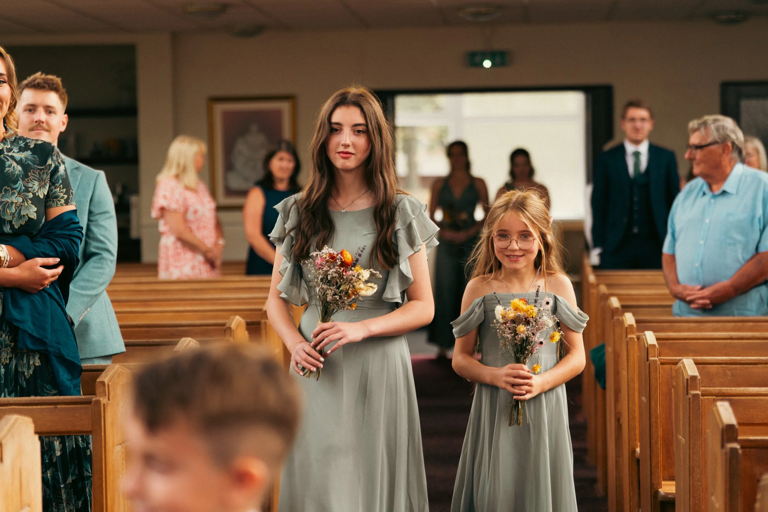 Two young girls in matching light green dresses holding bouquets of flowers walking down the aisle during a wedding ceremony in a church with guests seated on wooden pews.
