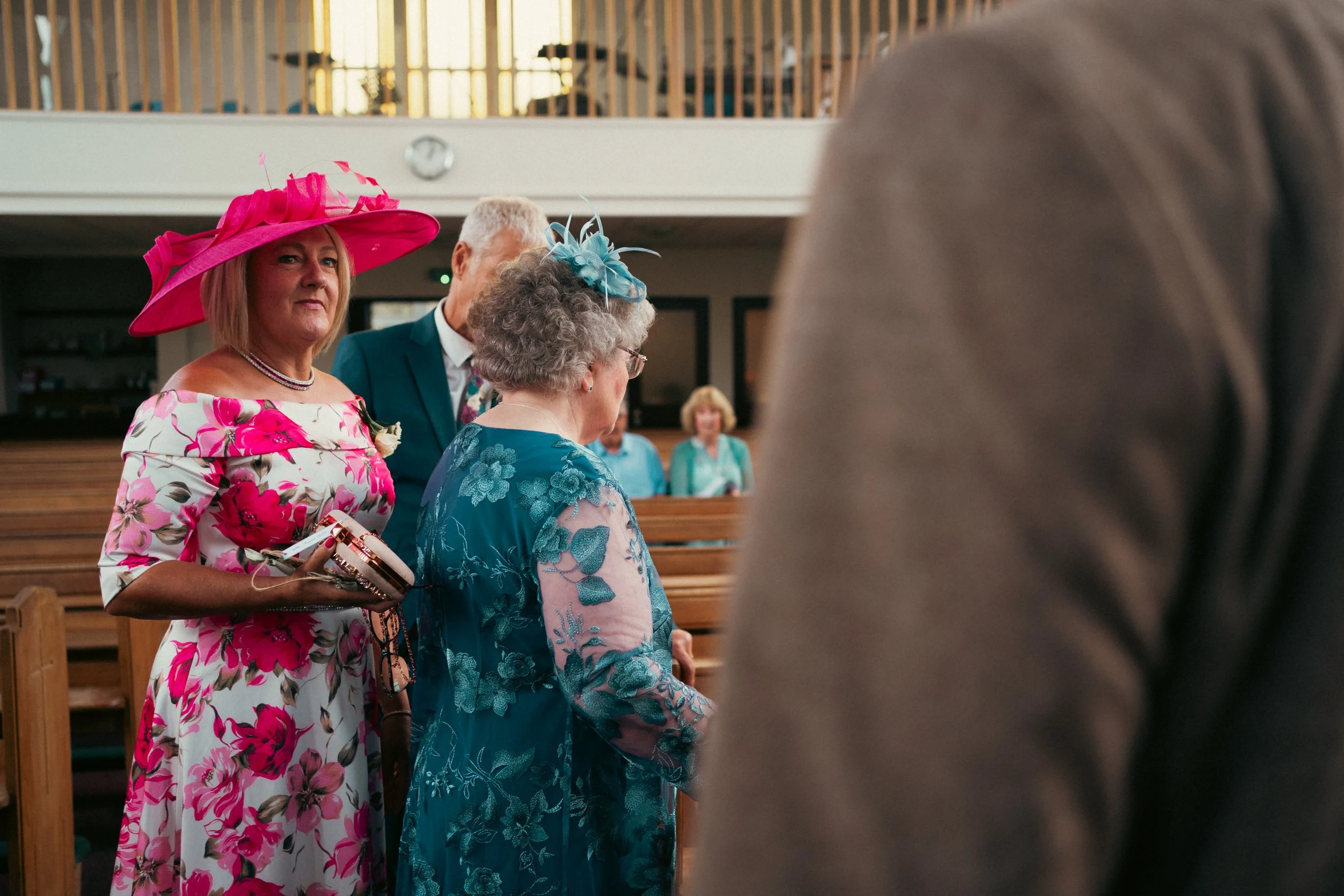 Group of elderly women and a man at a church service, dressed in colorful attire with hats, with pews and a few seated women in the background.