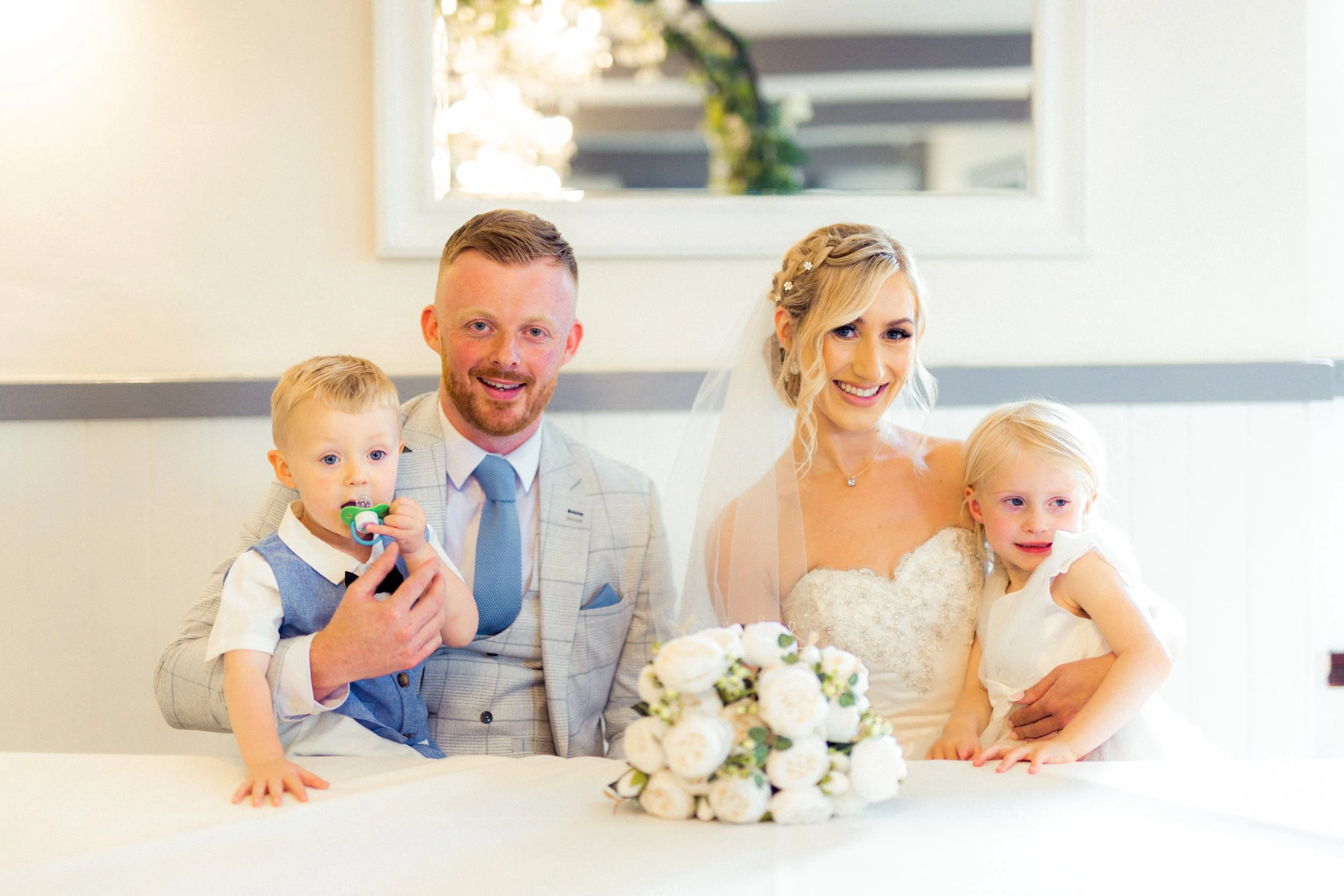 A bride and groom with two young children at a wedding reception, sitting at a table with a white floral centerpiece in front of them.