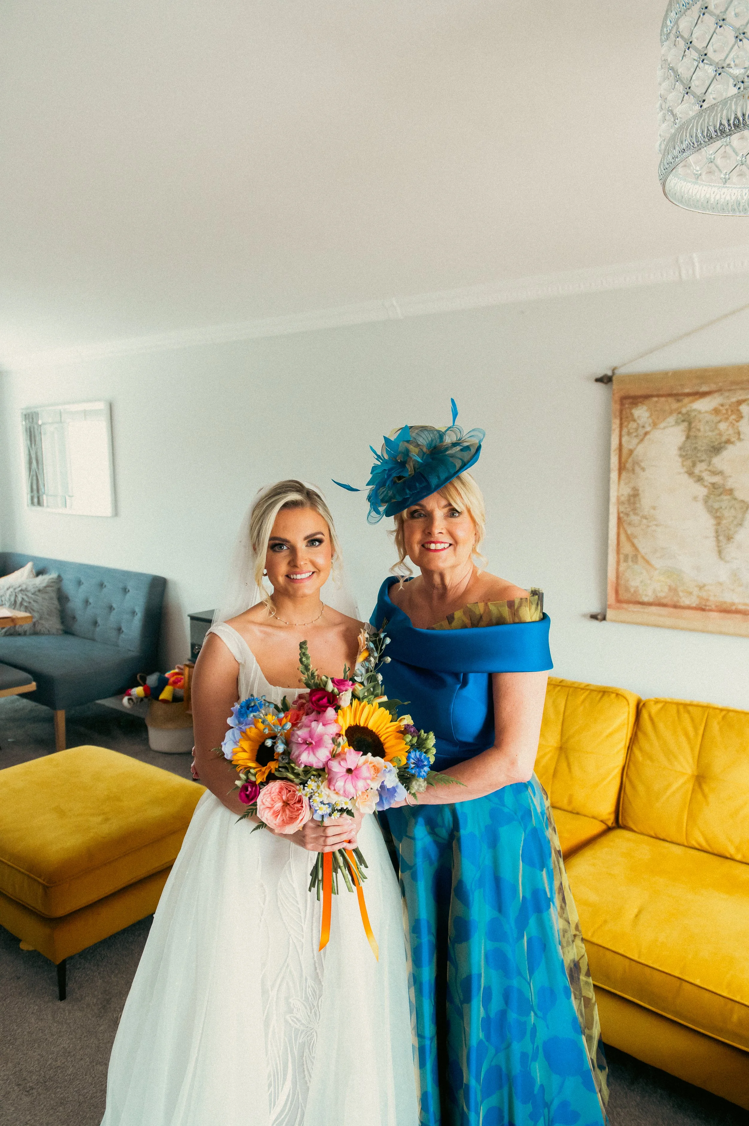 A bride in a white wedding dress holding a colorful bouquet of flowers standing next to an older woman in a vibrant blue outfit and matching hat inside a living room with a yellow couch and a world map on the wall.