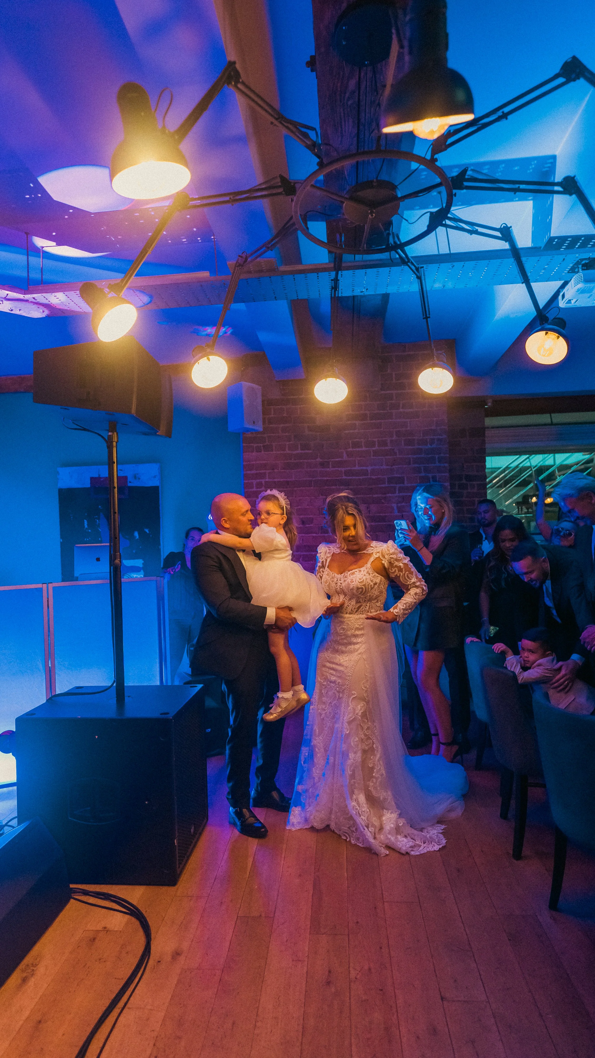 A group of people celebrating at a wedding reception, with a bride in a white lace gown and a groom in a dark suit, as a young girl in a white dress is held by a man. Guests are taking photos and interacting around them, with blue and purple lighting