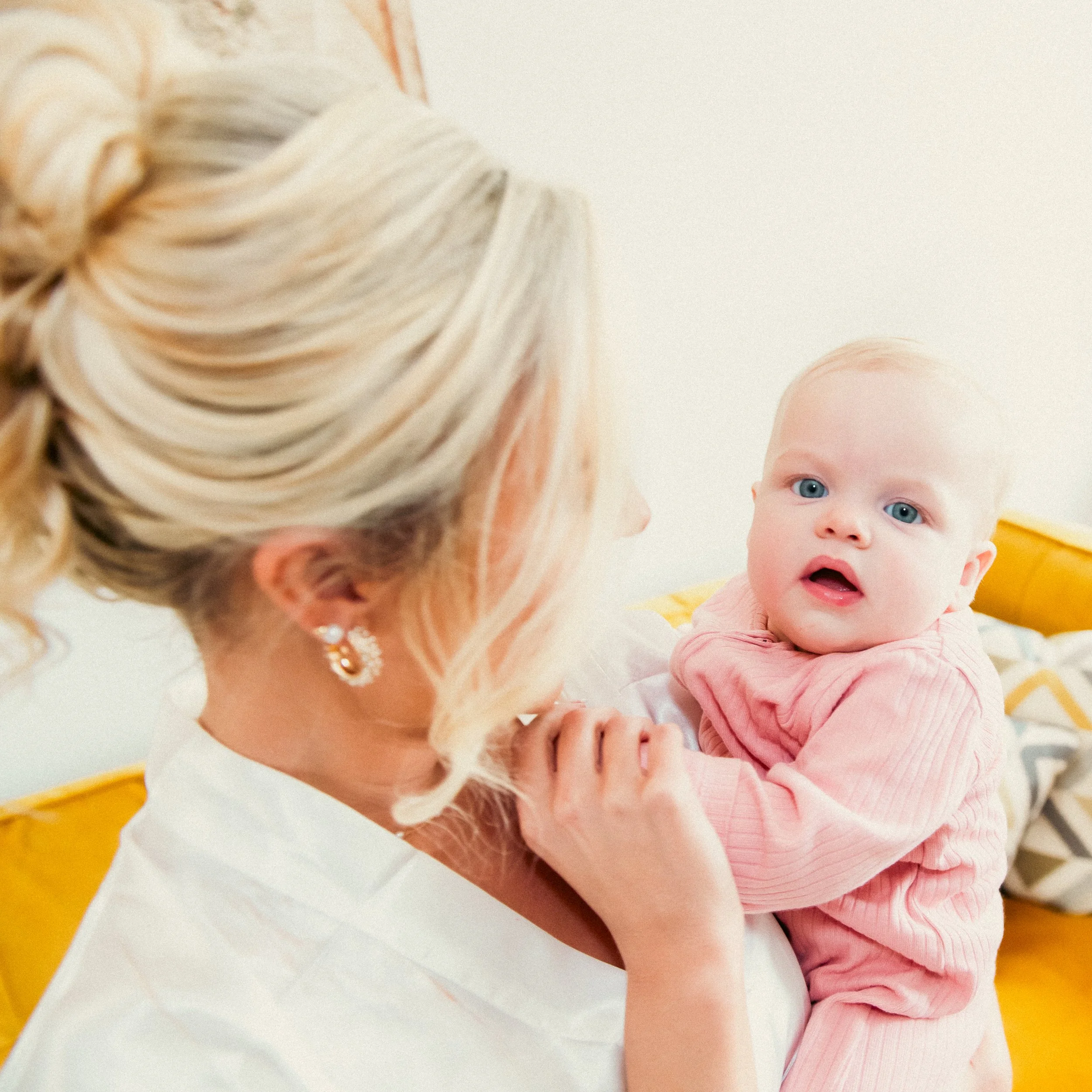 A woman with blonde hair and earrings holding a baby with blue eyes wearing a pink outfit, inside a home with a yellow couch and patterned pillows.