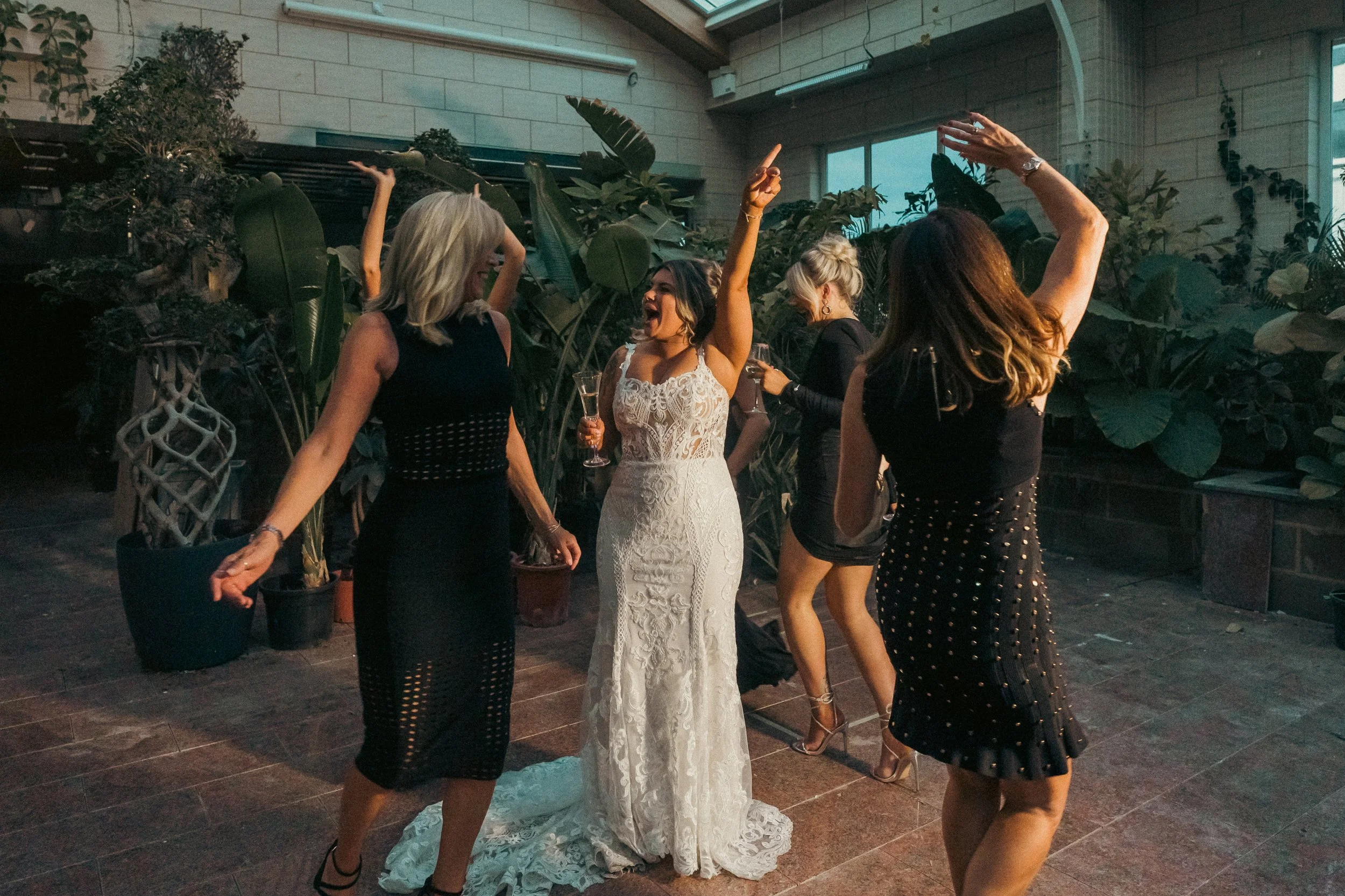 Women dancing and celebrating at a party, with one woman in a white lace wedding dress holding a glass of champagne.