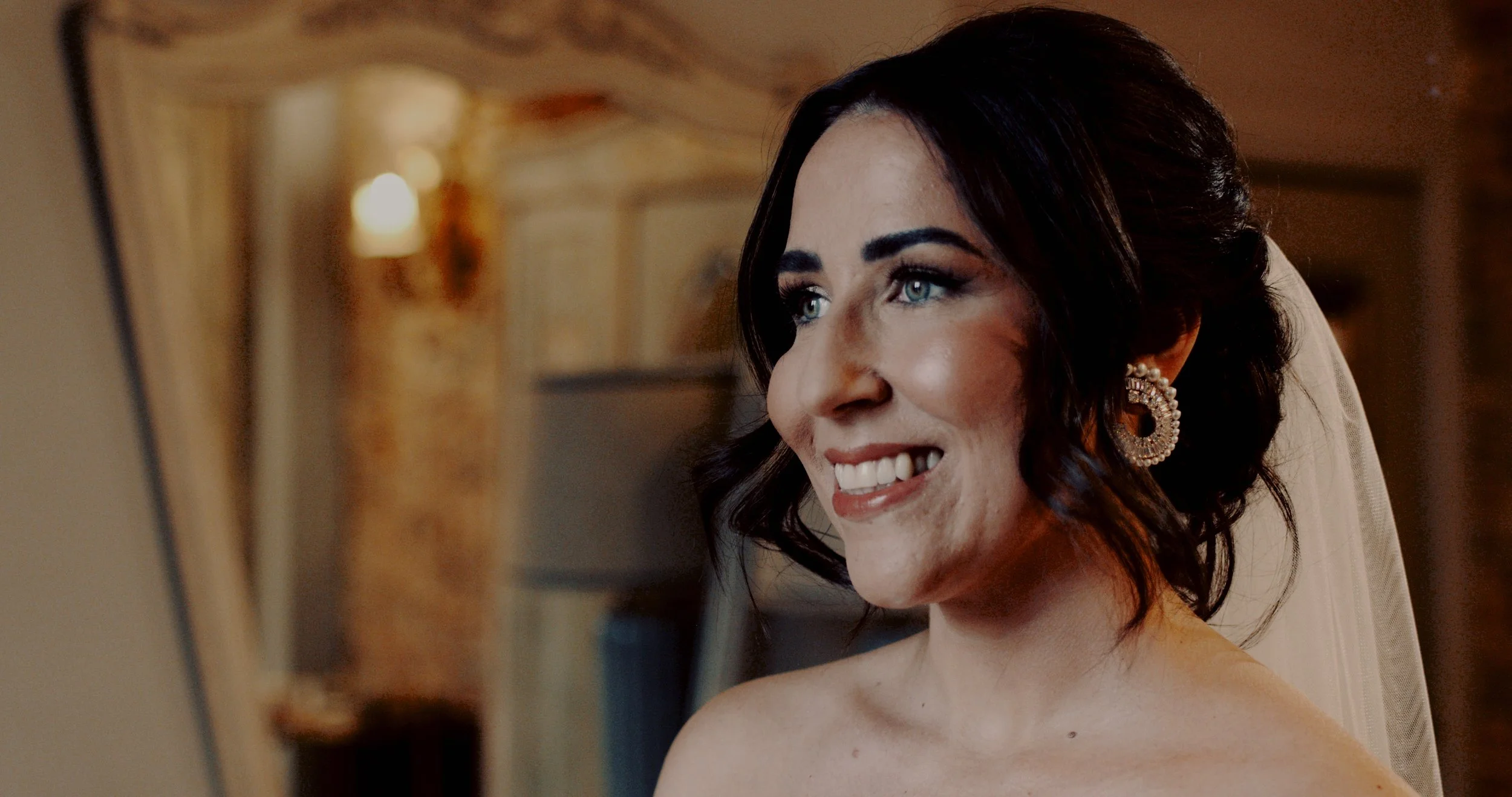 A woman with dark hair styled in loose curls, wearing pearl earrings and a wedding veil, smiling while looking at a mirror.
