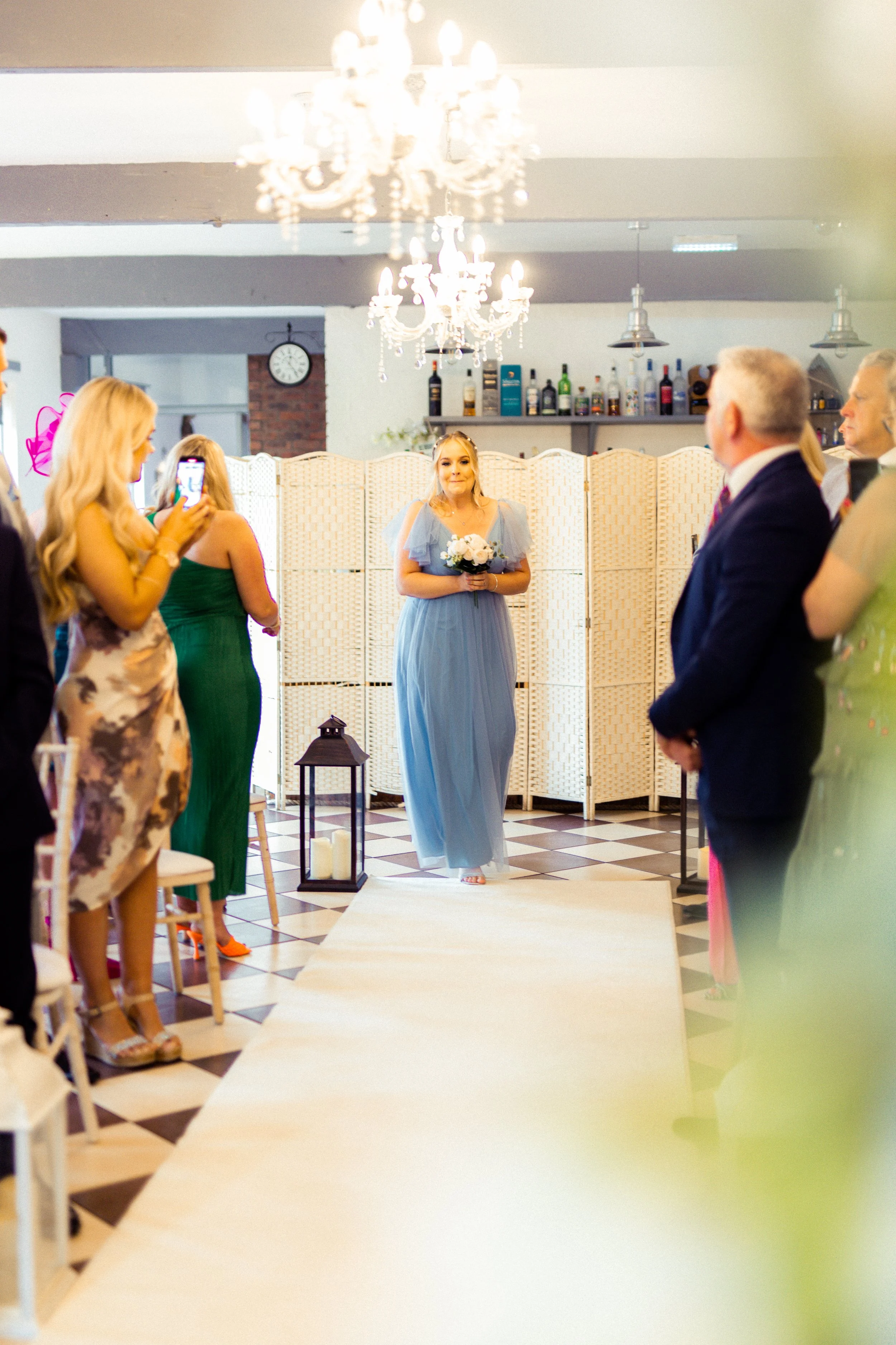 A bride walking down the aisle during her wedding ceremony, holding a bouquet, with guests standing on either side.
