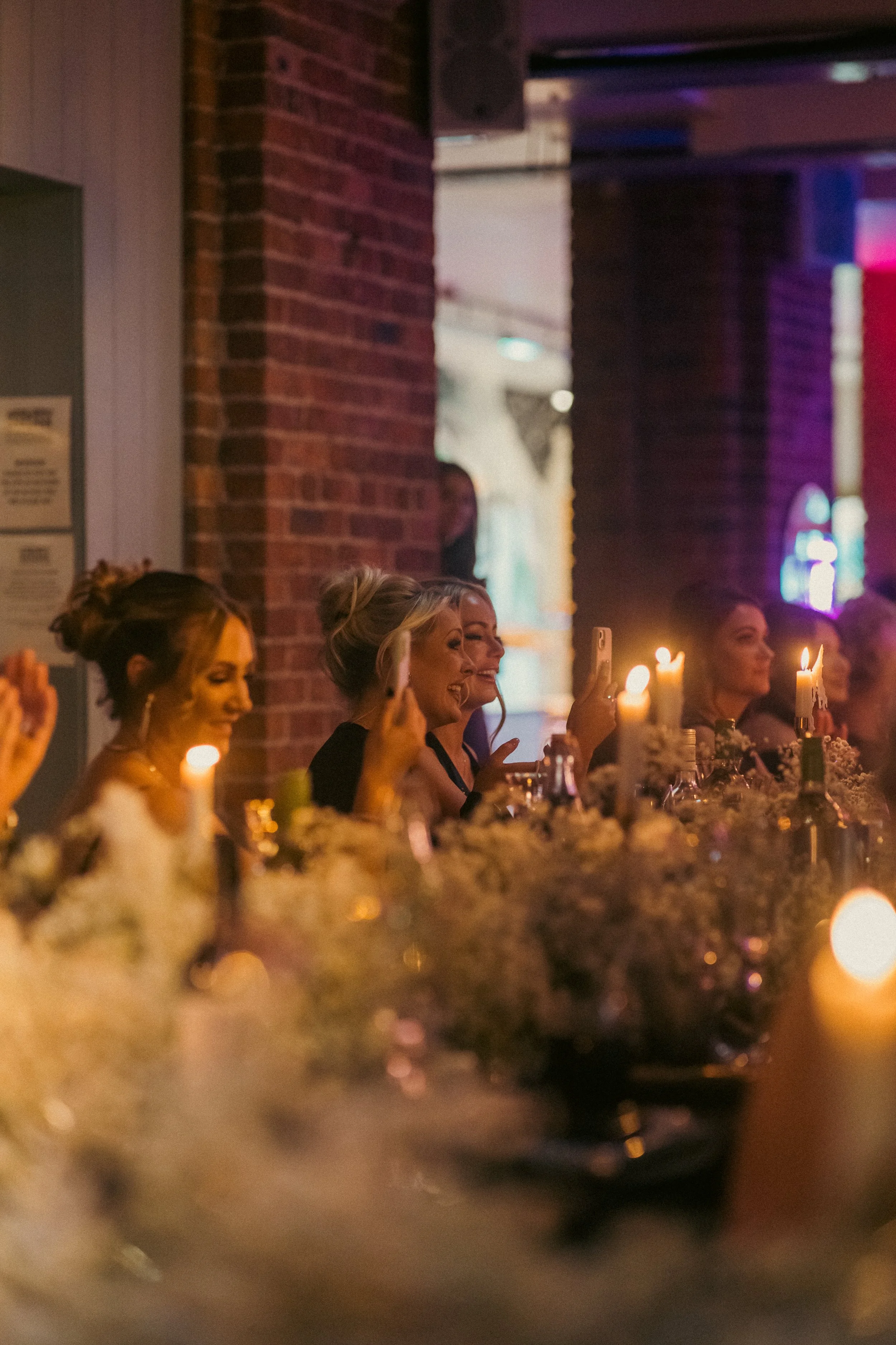 People sitting at a dimly lit banquet table, some taking photos, with candles and floral decorations, in an indoor venue with exposed brick walls.