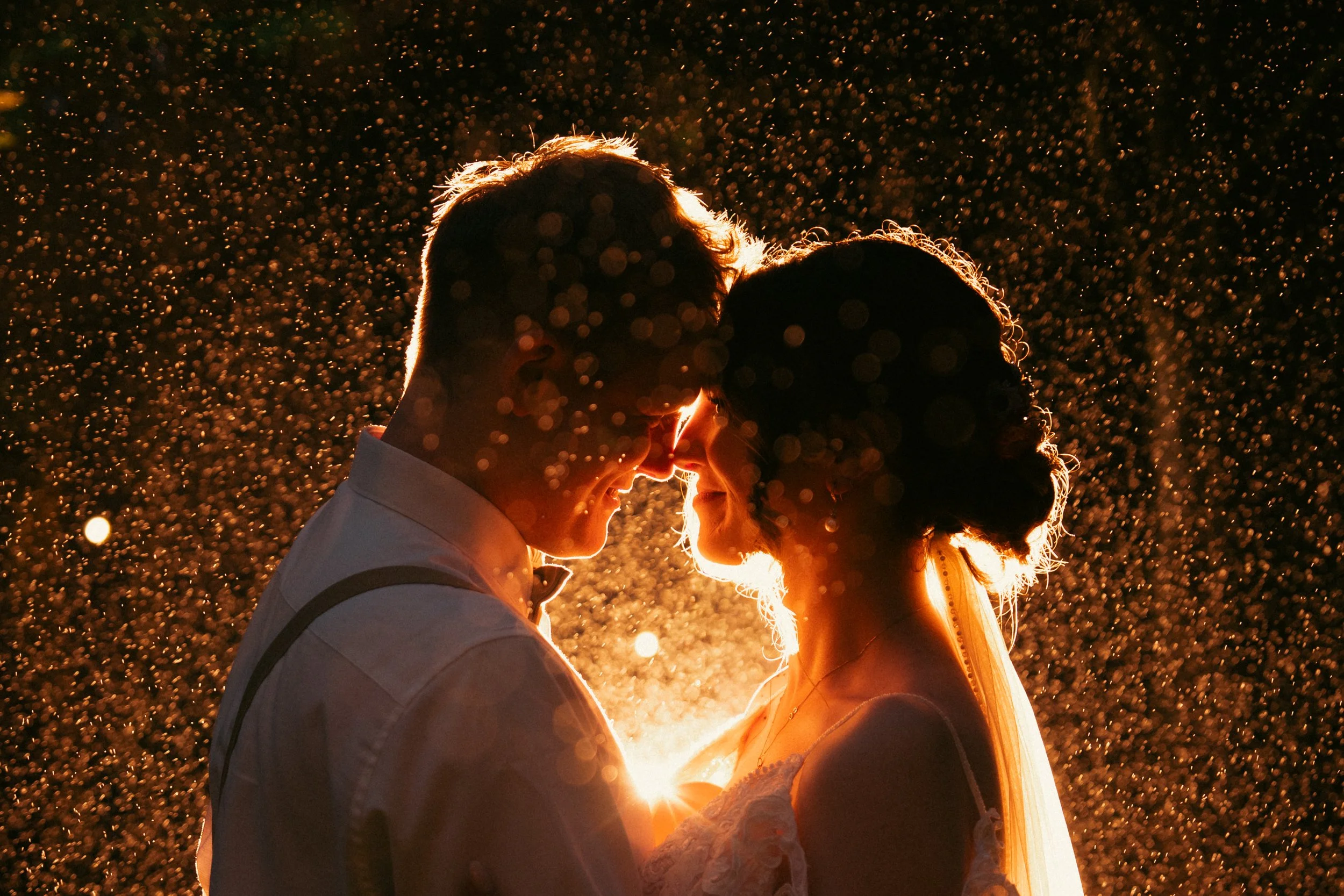 A couple faces each other closely at night, backlit by a warm glow, with falling rain or snow creating a sparkling effect around them.