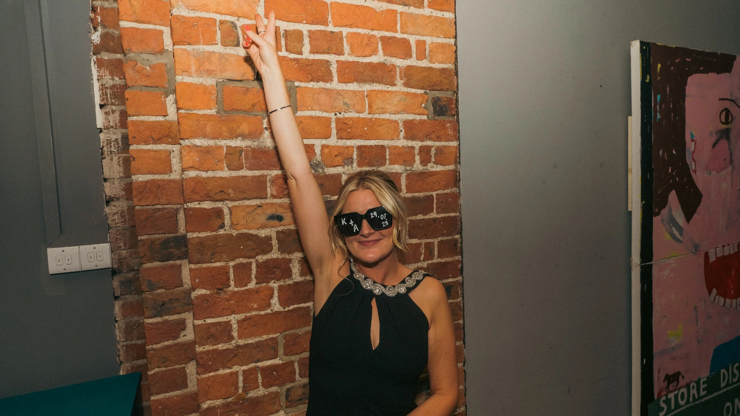 A woman with blonde hair wearing black sunglasses with white writing, a black sleeveless dress with a decorative collar, sitting in front of a brick wall. She is smiling and raising her right arm high, making a peace sign.