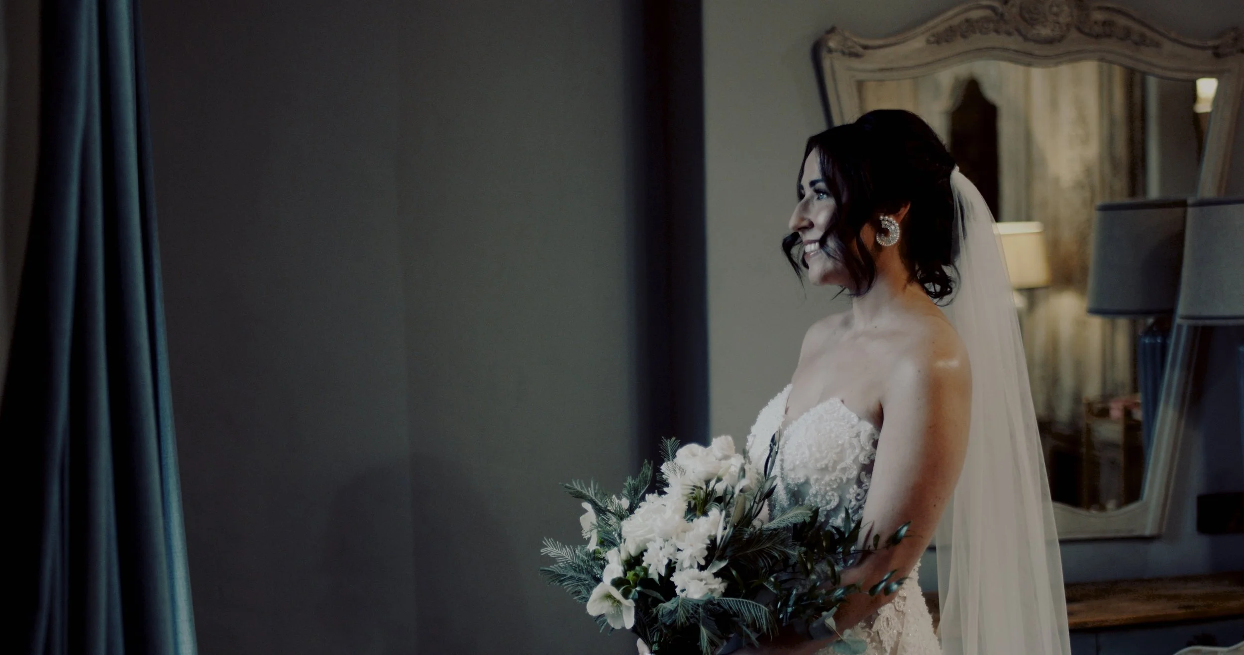 Bride in a strapless lace wedding gown holding a bouquet of white flowers and greenery, smiling, with a vanity mirror and lamps in the background.