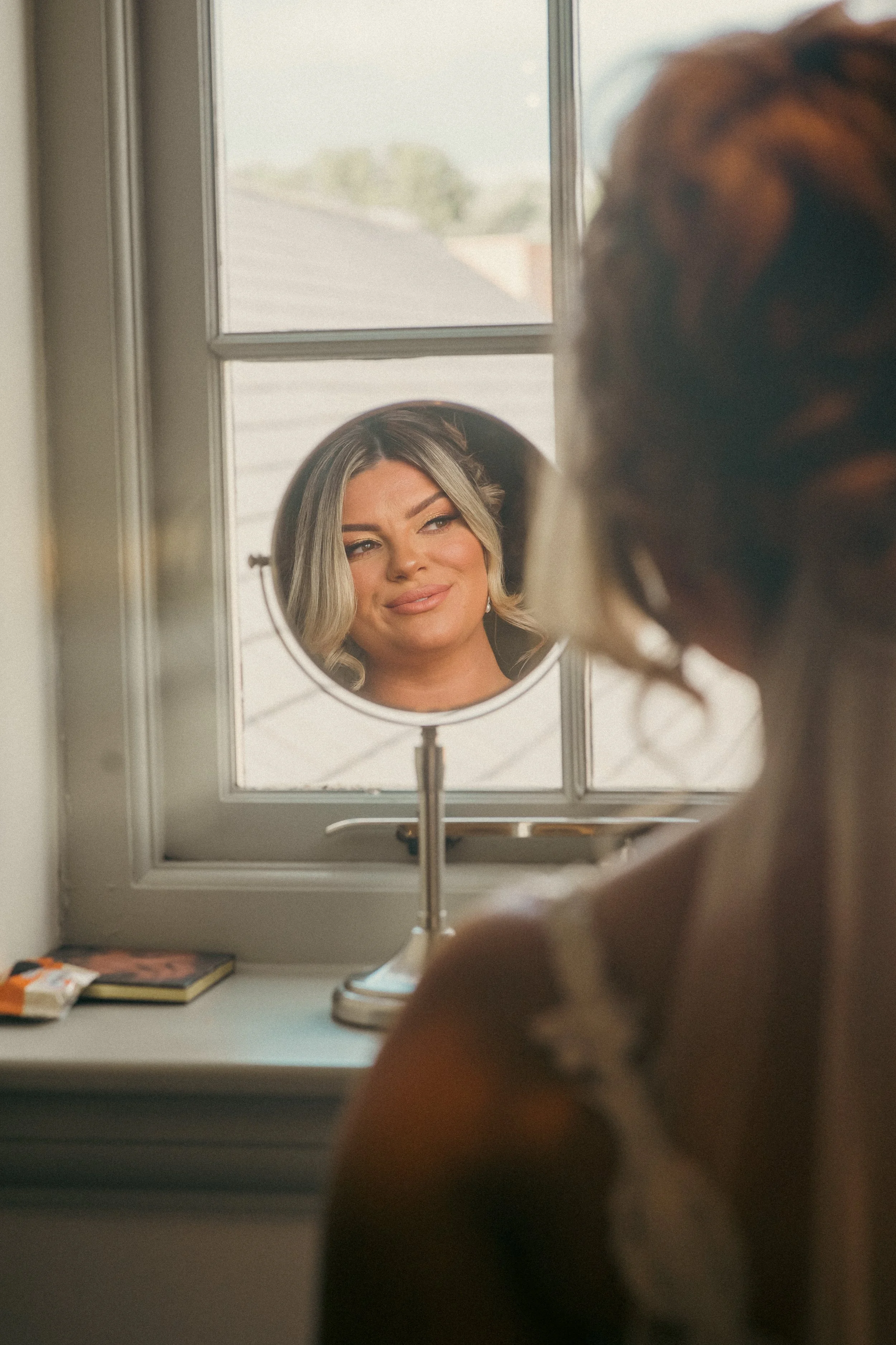A woman gazing into a small round mirror on a window sill, with her reflection showing a subtle smile and makeup, in a well-lit room with a book and snack on the window sill.