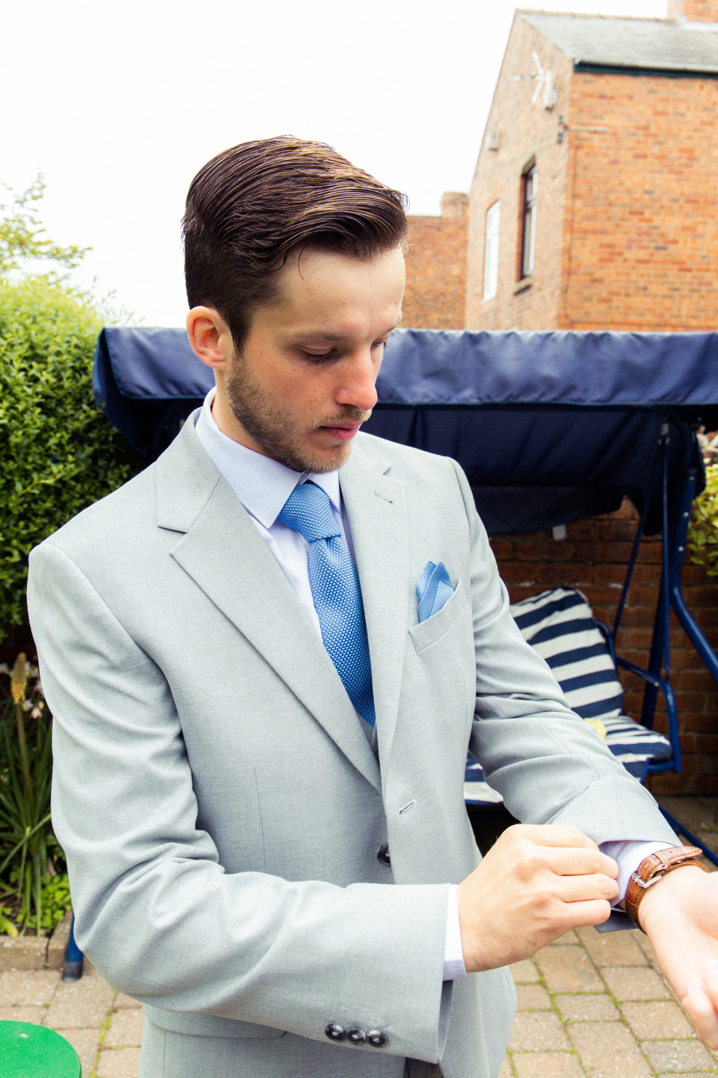 A man in a light gray suit and blue tie adjusts his watch outdoors in a garden setting, with a brick house and a garden swing in the background.