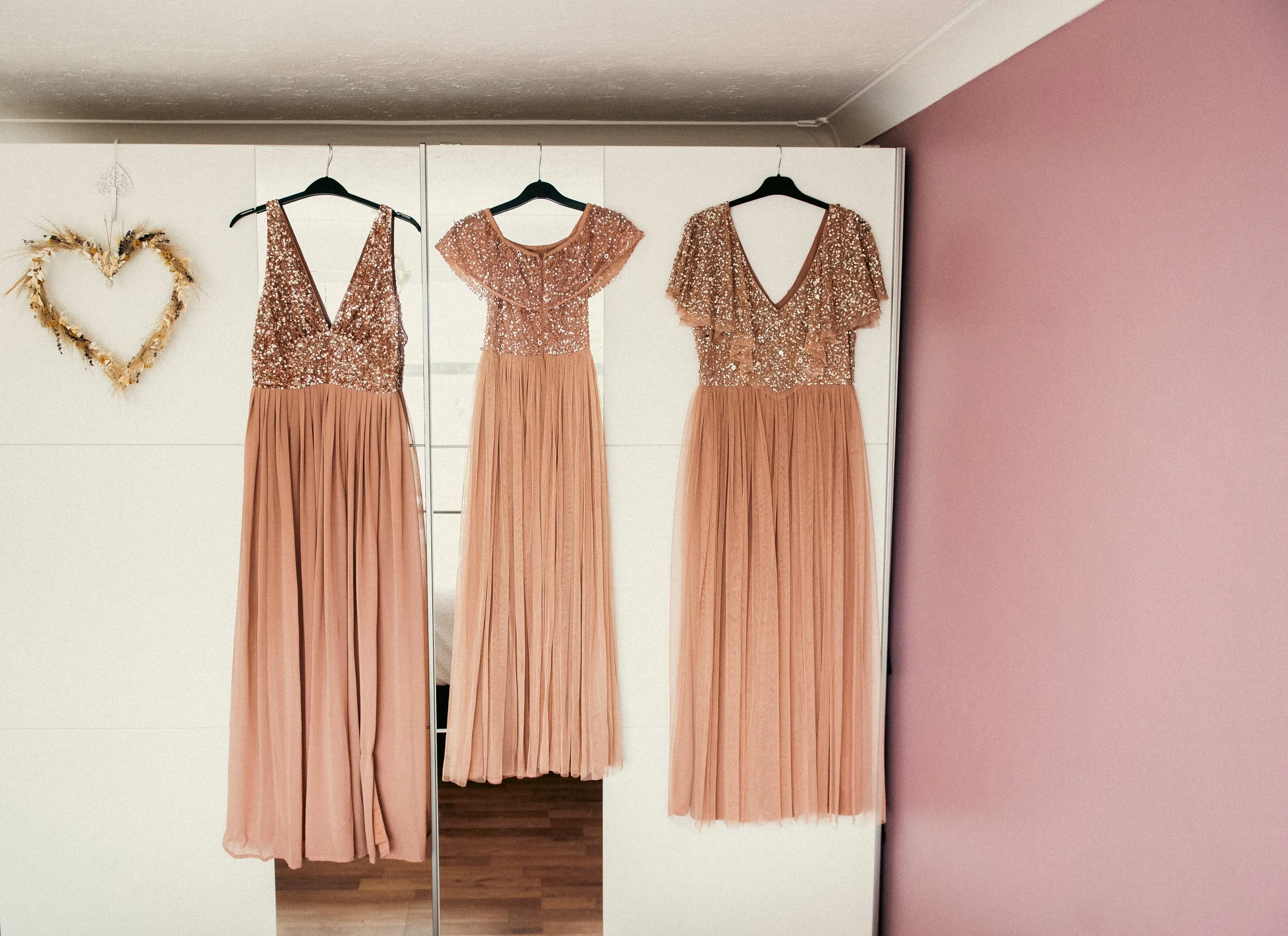 Three peach-colored formal dresses with sequined bodices and pleated skirts hanging on black hangers in front of a white wardrobe and a pink wall, with a heart-shaped wreath made of dried flowers on the wall to the left.