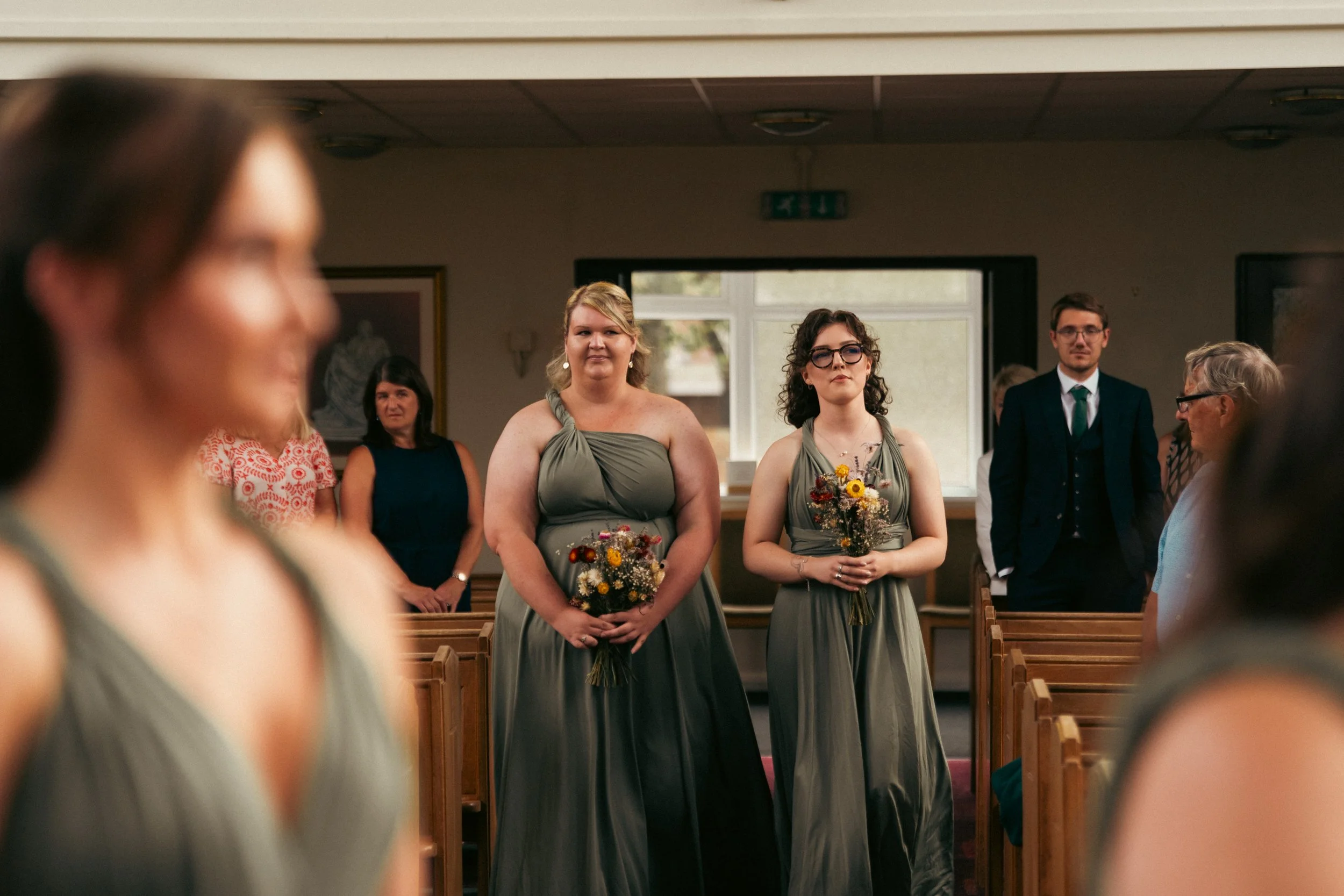 Two bridesmaids in matching green dresses standing in a chapel, holding small bouquets of flowers during a wedding ceremony.