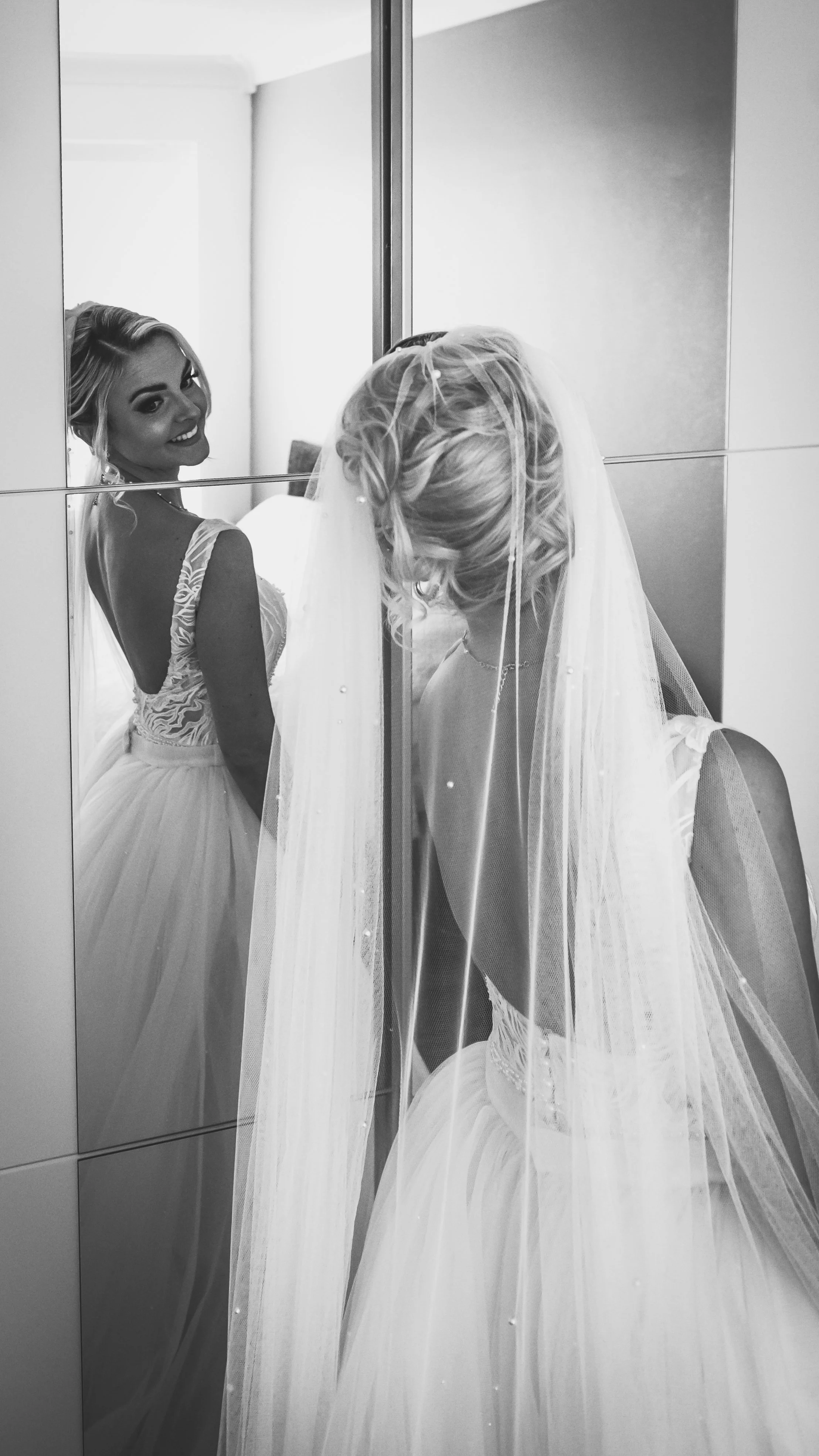 A bride in a wedding dress and veil stands in front of a mirror, smiling at her reflection, with her reflection also visible in the mirror.